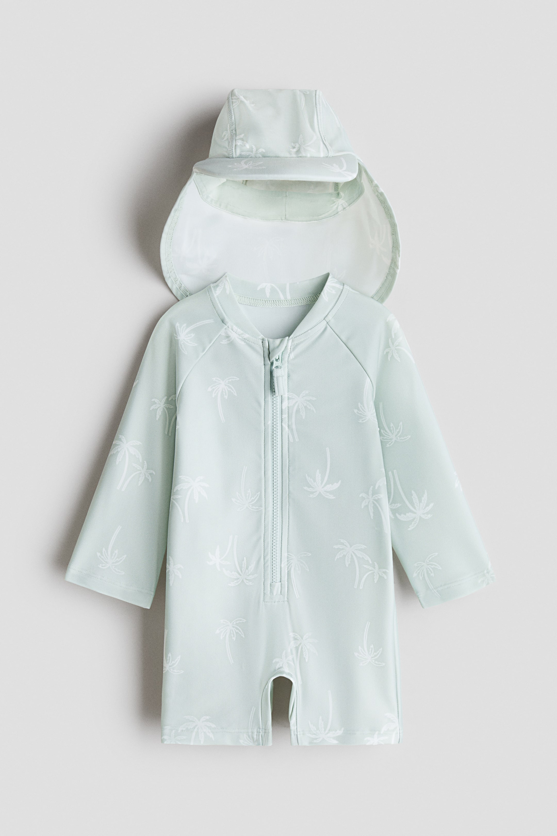 View larger image: A light green sun protection suit and matching cap display a white palm tree pattern. The all-in-one suit, shown from the front, features a small stand-up collar, long raglan sleeves, short legs and a full-length front zipper with a chin guard, while the cap has a front peak and a rear flap.