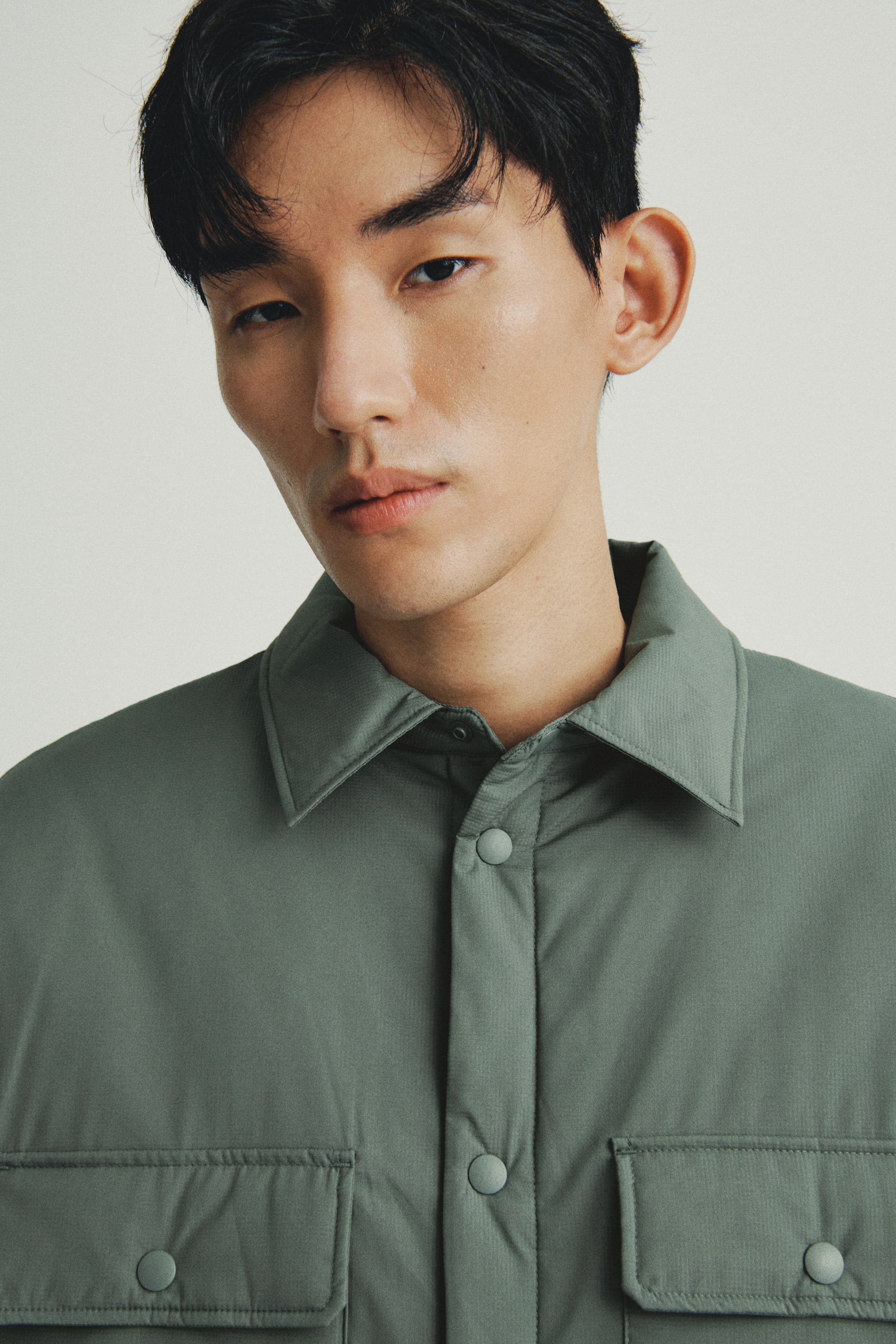 이미지 확대 보기: 어두운 톤의 muted 녹색 오버셔츠는 뾰족한 칼라와 스냅 버튼이 있는 앞 플래킷, 그리고 각 스냅 버튼으로 잠글 수 있는 플랩이 있는 두 개의 가슴 포켓이 특징입니다. 이 의상은 약간 패딩 처리가 되었으며 섬세한 텍스처 마감이 돋보입니다.