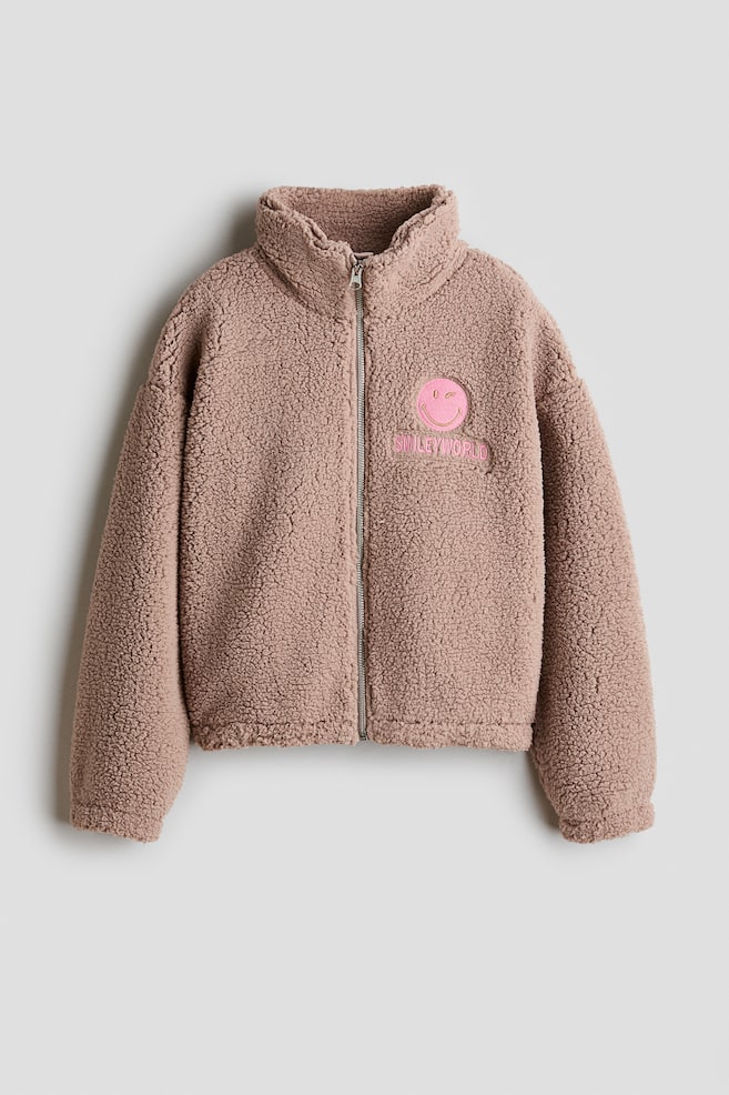 A dusty pink, loose-fit teddy jacket is shown from the front, featuring a high stand collar, a central zip, dropped shoulders, and elasticated cuffs and hem. The jacket is detailed with a pink winking smiley-face patch and the embroidered text "SMILEYWORLD" on the left chest. - 1