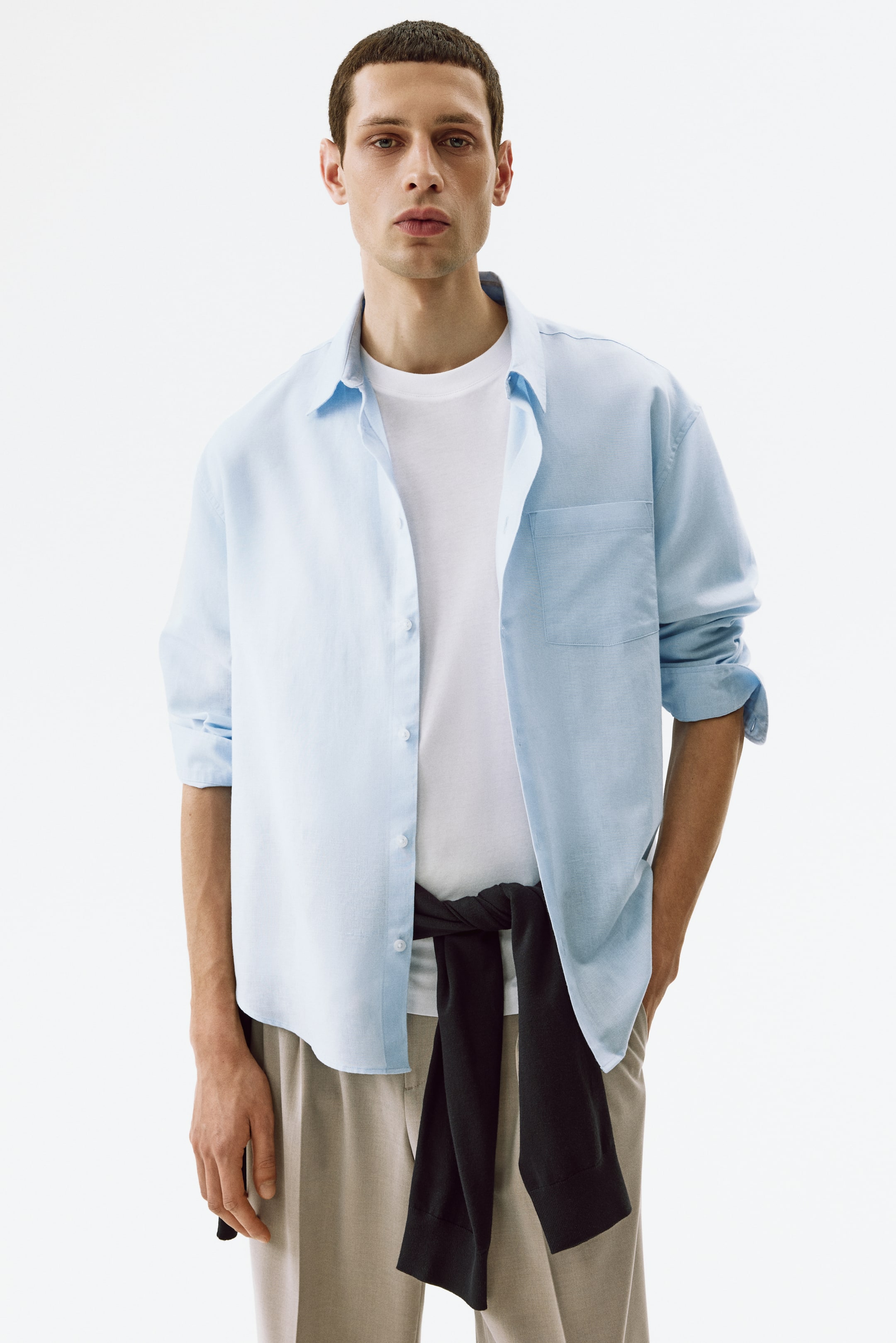 Größeres Bild ansehen: Ein hellblaues Hemd mit Knopfleiste ist nach vorne gerichtet und wird offen über einem weißen T-Shirt getragen. Es verfügt über einen klassischen Kragen, eine aufgesetzte Brusttasche links und hochgekrempelte Ärmel. Ein schwarzer Pullover ist über dem Hemd um die Taille gebunden.