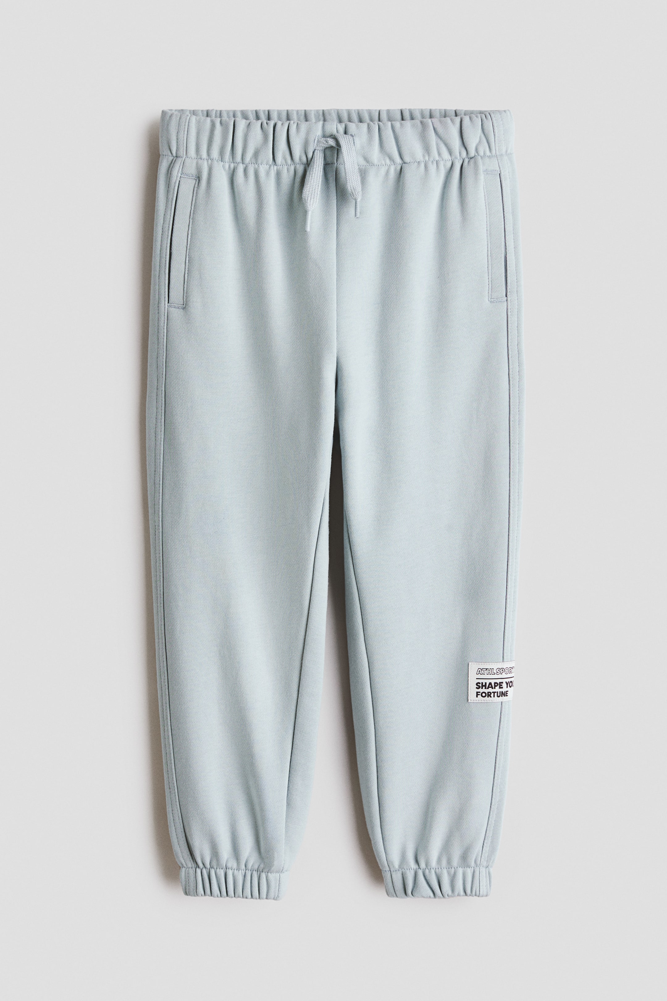View larger image: Light blue sweatpants feature an elasticated waistband with a drawstring, two side slash pockets, and ribbed ankle cuffs. A white rectangular patch with the text 'ATHL. SPORT SHAPE YOUR FORTUNE' is stitched onto the lower right leg.