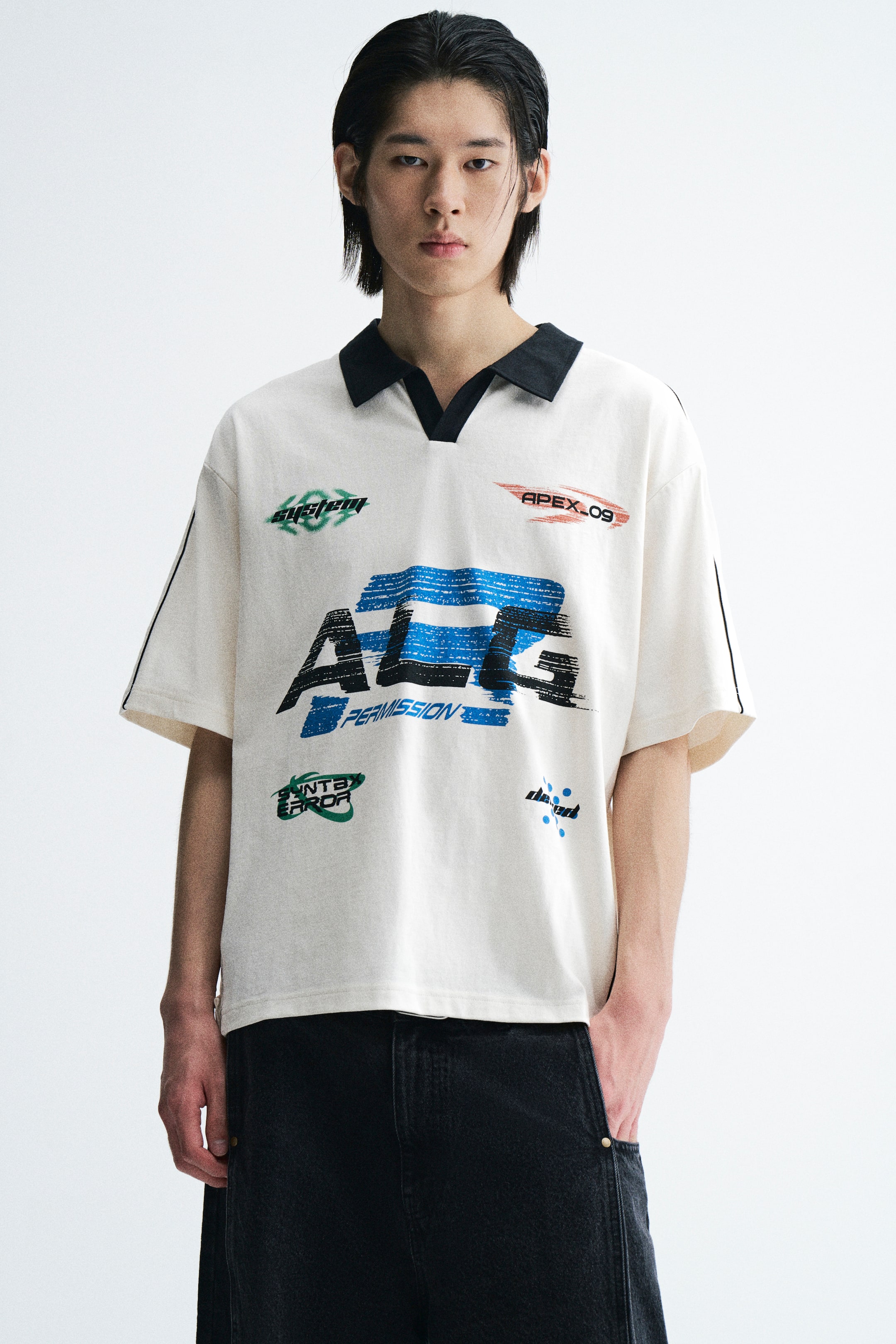 View larger image: A loose-fit, off-white short-sleeve polo shirt with a contrasting black open collar and multiple graphic prints is worn by the model. The shirt features dropped shoulders, thin black piping along the sleeves, and a straight hem, paired with dark trousers.