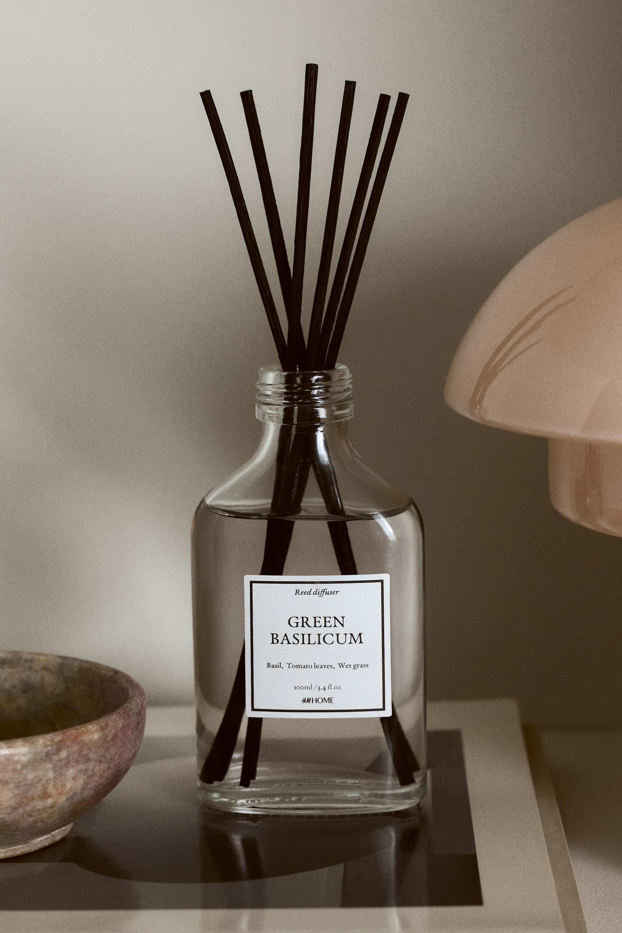 Näytä suurempi kuva: Edestäpäin kuvattu tuoksutikkuasetti koostuu kirkkaasta, suorakulmaisesta lasipullosta, joka on täytetty vaaleanvärisellä nesteellä. Useita tummia bambutikkuja on upotettu nesteeseen ja ne ulottuvat pullosta ylöspäin. Pullon etuosassa on valkoinen etiketti, jossa lukee "GREEN BASILICUM" ja tilavuus "100 ml / 3.4 fl.oz".