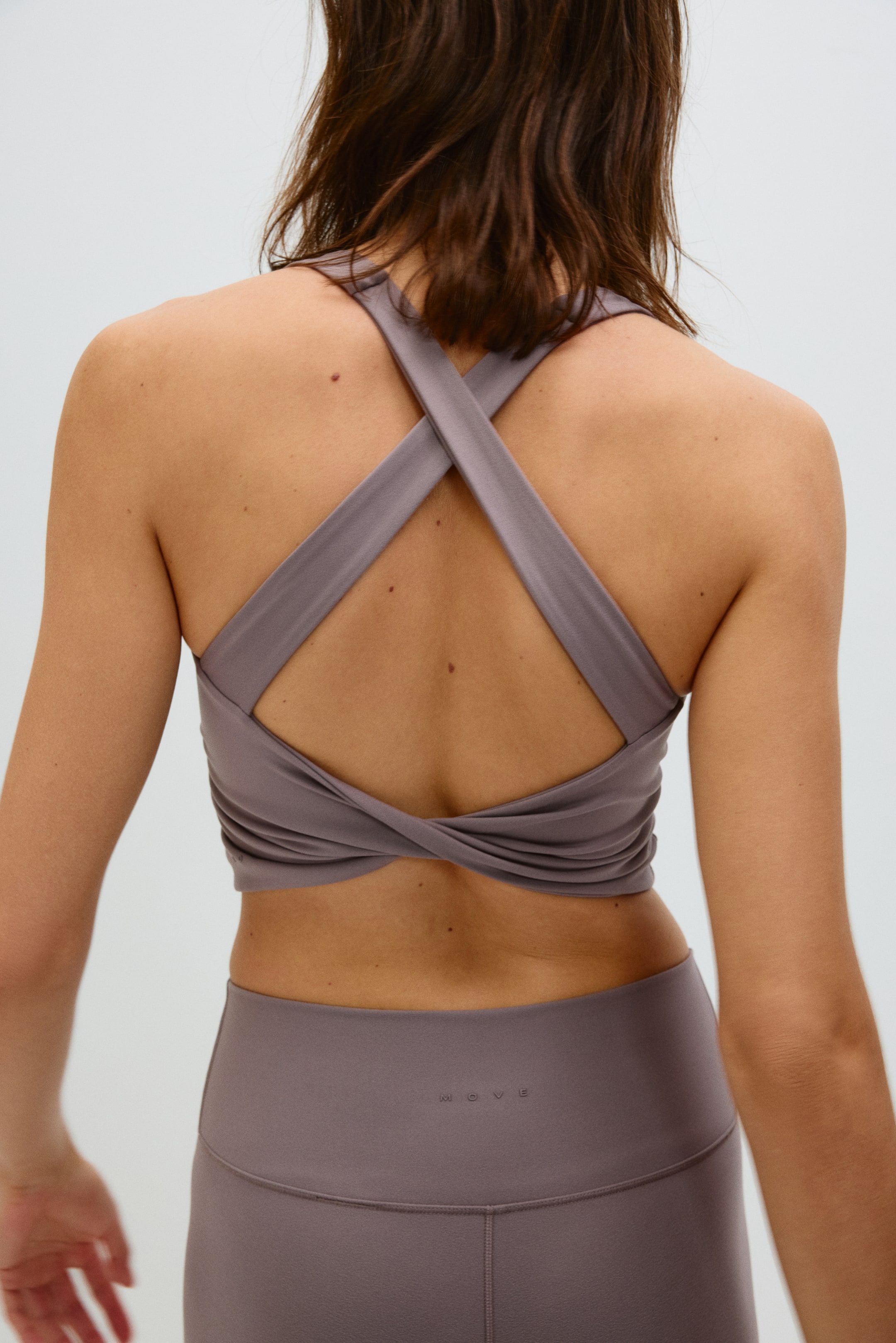Ver imagen más grande: Se presenta un top corto atlético color malva y leggings de cintura alta a juego desde la parte trasera. El top corto cuenta con tirantes anchos cruzados que se retuercen en la parte baja de la espalda, y los leggings muestran la palabra 'MOVE' en relieve en la cintura trasera.