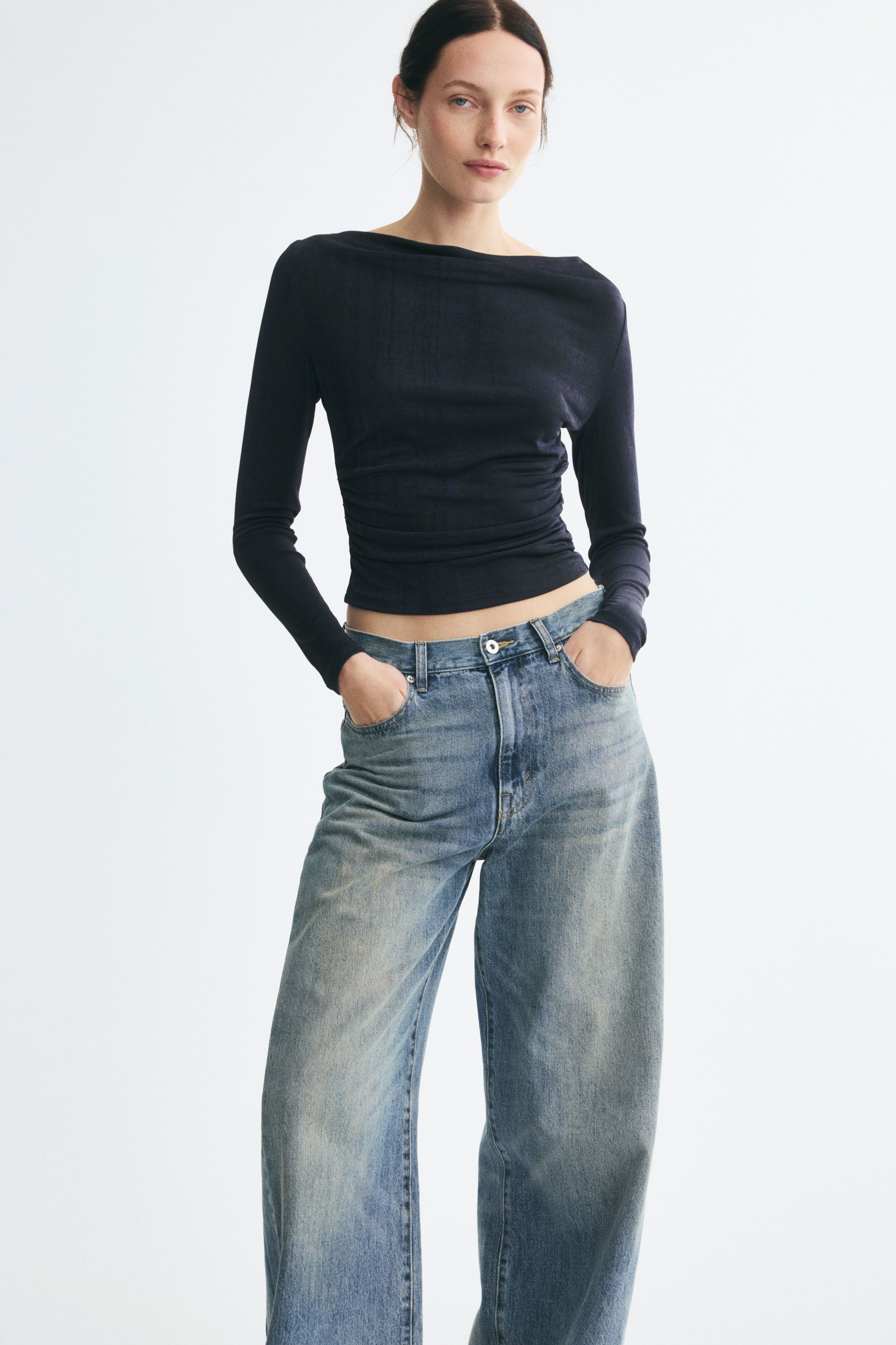 A black long-sleeved top facing front showcases a wide boat neckline and a textured, slightly lustrous fabric. It features ruched detailing along the lower side seams, creating draped horizontal folds across the front.