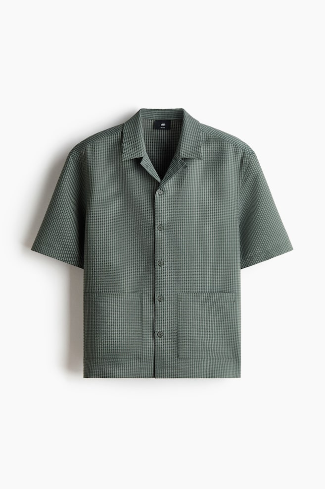 A dark green short-sleeved shirt is showcased front-facing, boasting a waffle-knit texture that creates a subtle grid pattern. It features a Cuban collar, a full button-front closure, and two patch pockets on the lower front. - 1