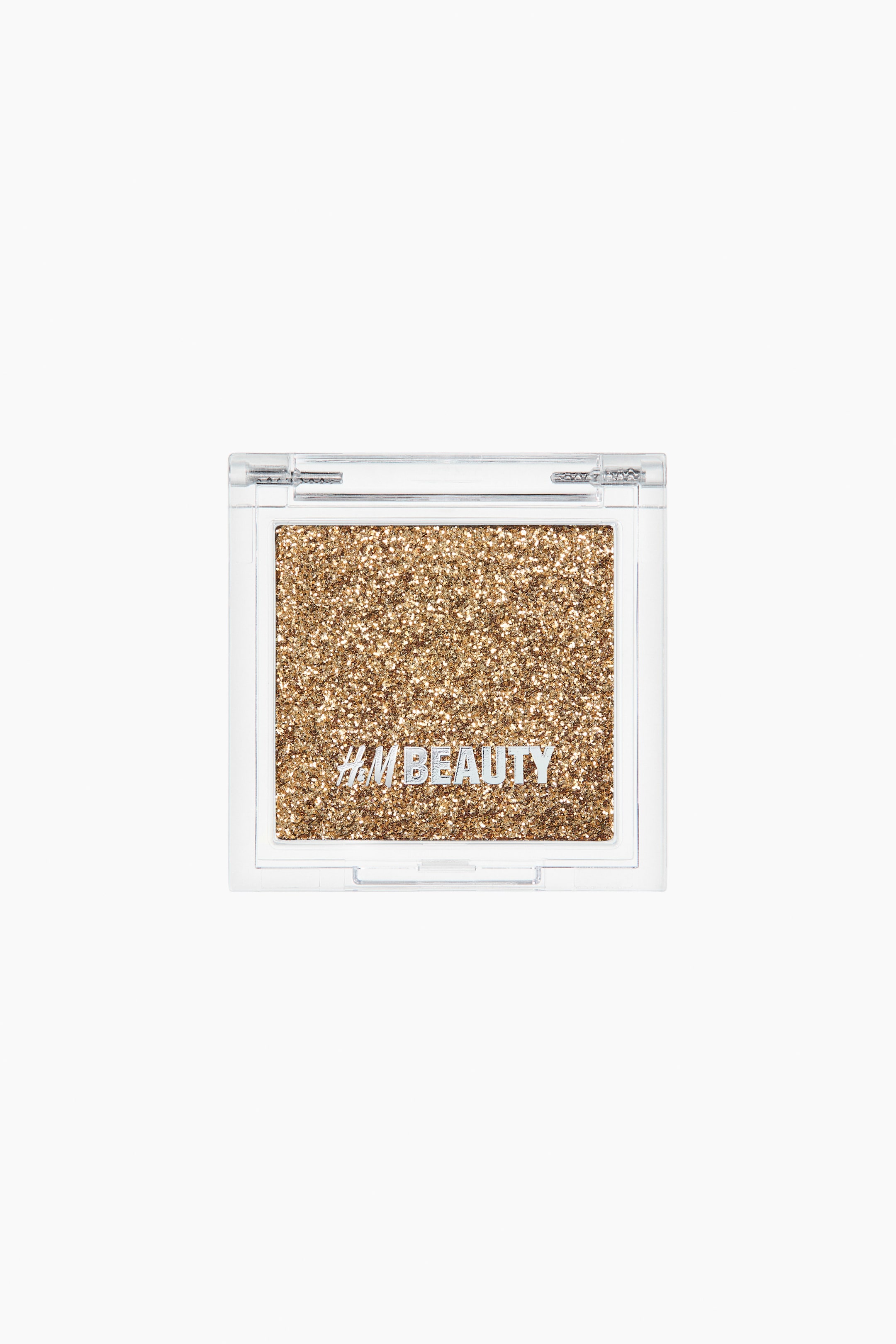 Zobacz większy obraz: Cień do powiek. Prostokątna paleta złotego, brokatowego cienia do powiek zamknięta jest w przezroczystym plastikowym opakowaniu, skierowanym przodem. Na brokatowej powierzchni widnieje biały napis "H&M BEAUTY".
