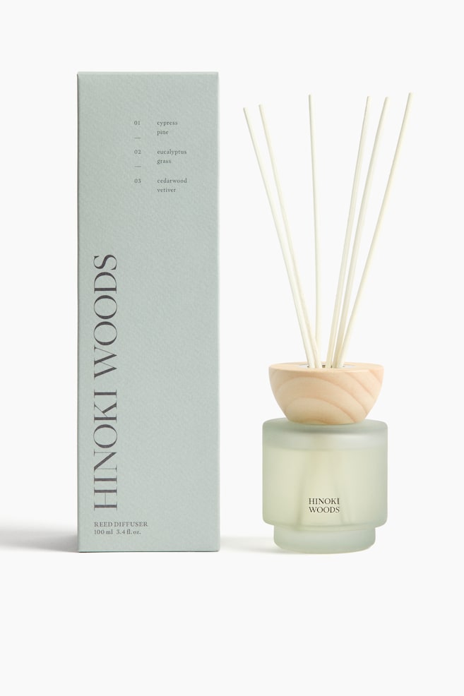 A frosted glass reed diffuser bottle is standing, featuring a distinctive stepped base and the text "HINOKI WOODS" printed on its front. It is filled with a light-coloured liquid and capped by a light wooden collar, from which several wooden reeds are extending vertically. A coordinating rectangular box is standing upright to the left, displaying product branding and scent details. - 1