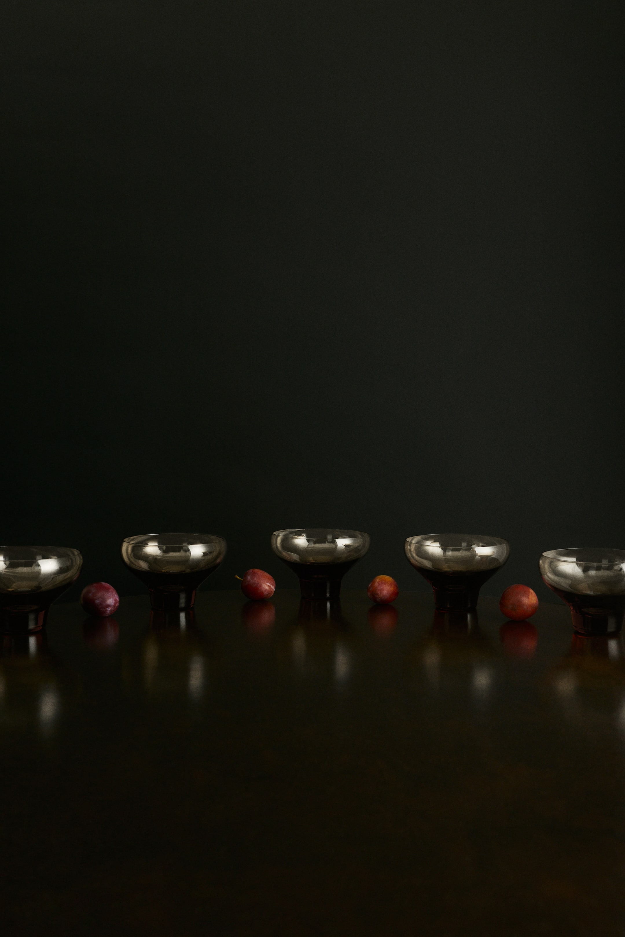 View larger image: Mouth-blown glass bowls are arranged in a horizontal row, facing forward, showcasing their wide, shallow, smoke-tinted vessels and thick, solid dark bases. Small round plums rest between the bowls on a dark reflective surface.