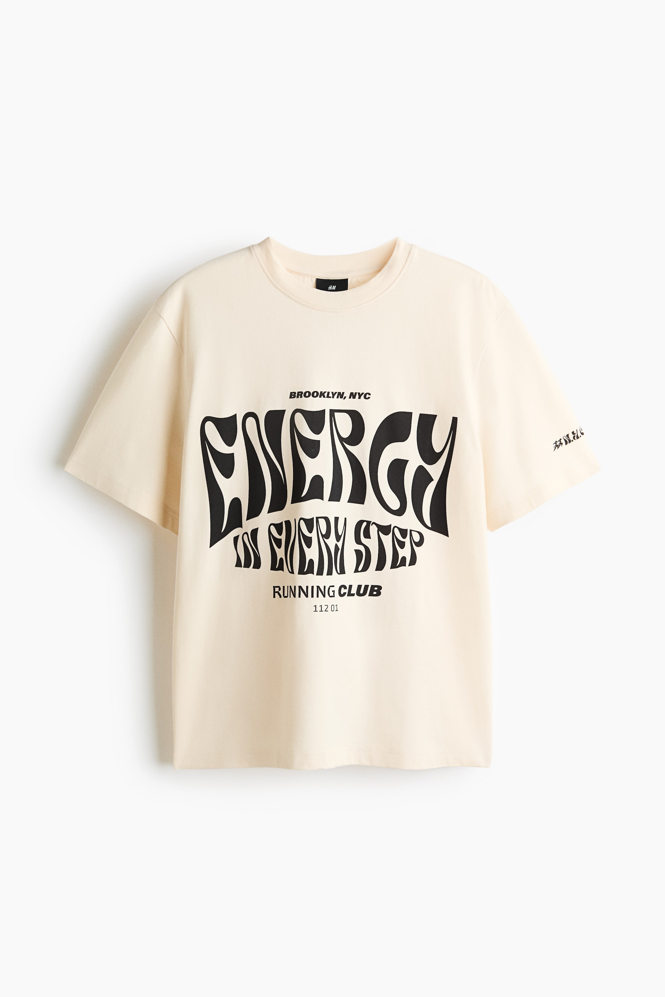 View larger image: A light beige T-shirt in midweight cotton jersey is shown from the front, featuring a round, rib-trimmed neckline, gently dropped shoulders, and a straight-cut hem. A large black print motif reading "ENERGY IN EVERY STEP," along with "BROOKLYN, NYC" and "RUNNING CLUB" details, is adorning the front, and stylised black script is appearing on the right sleeve.