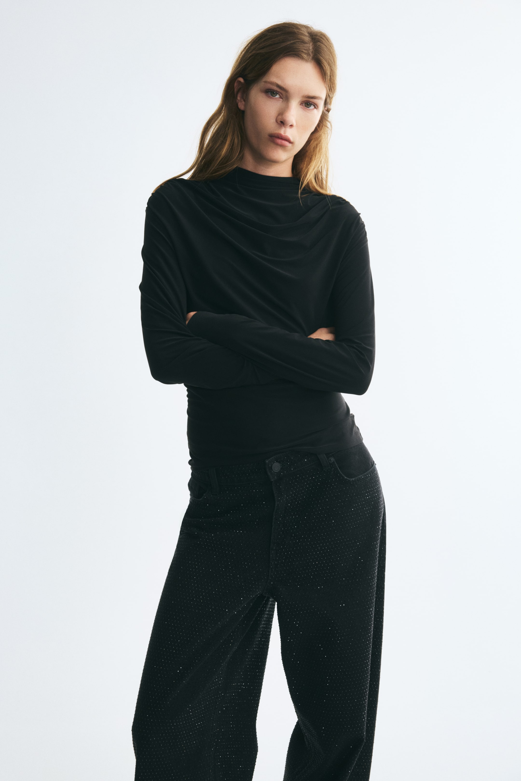 Agrandir l'image: Un haut noir ajusté à manches longues, doté d’un col montant et de fronces drapées sur la poitrine, est porté par le mannequin, qui se tient les bras croisés. Le haut est associé à un pantalon foncé à texture marquée.