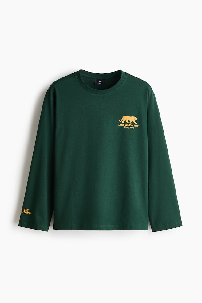 A dark green, long-sleeve T-shirt is hanging front-facing, showcasing a round, rib-trimmed neckline, dropped shoulders, and a straight-cut hem. It features a small yellow print on the left chest of a big cat silhouette with the words "Don't Let The Fear Stop You" underneath, along with the text "BE BRAVE" printed on the right sleeve cuff. - 1