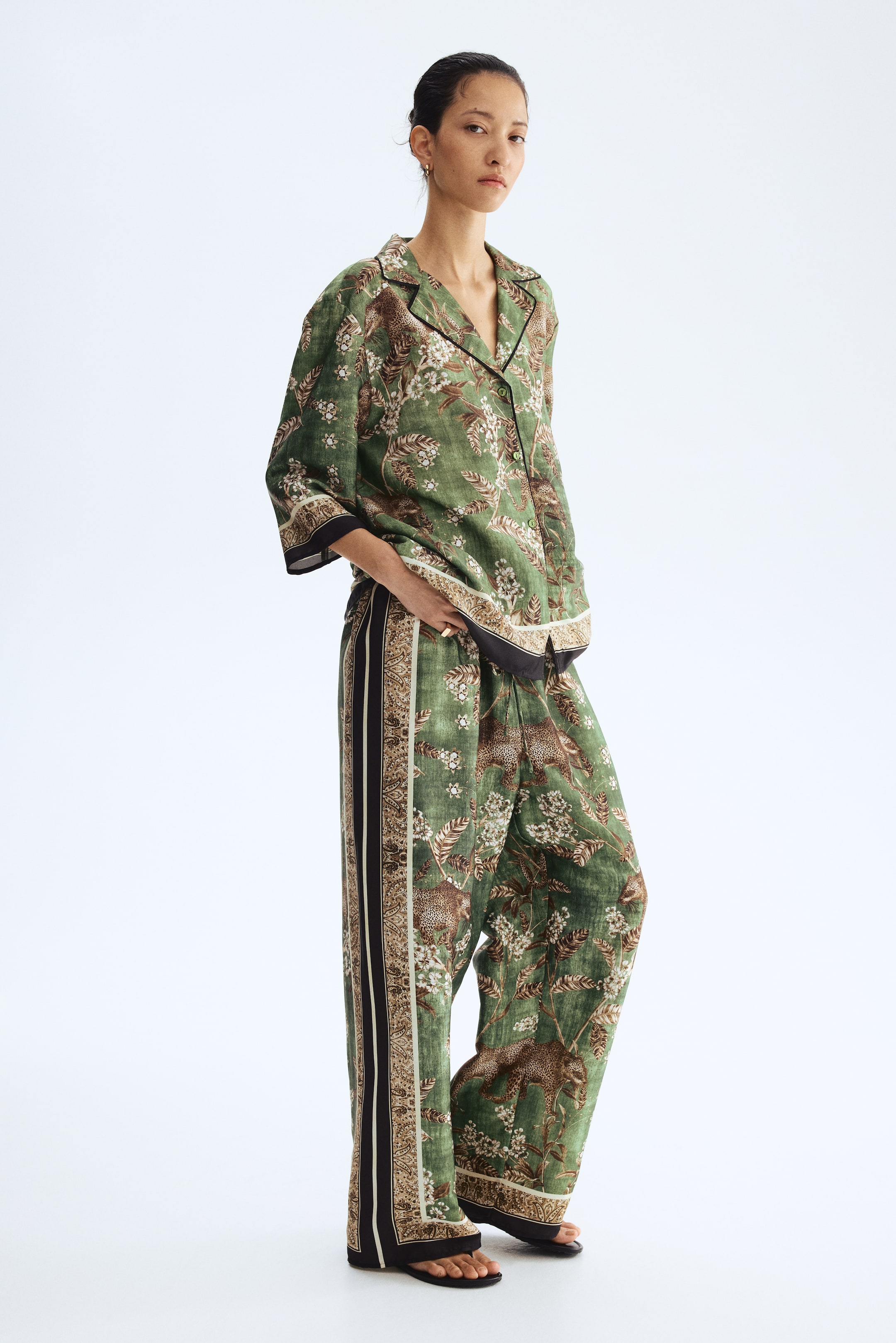 Ver imagen más grande: Un conjunto de camisa y pantalones de pierna ancha a juego en un patrón verde, con motivos de leopardos y flores, es llevado por la modelo. La camisa con cuello tiene mangas cortas y ribetes negros a lo largo de sus bordes, y los pantalones de pierna ancha incluyen una franja lateral con un patrón destacado. La modelo está colocando una mano en el bolsillo del pantalón y también lleva sandalias planas negras.