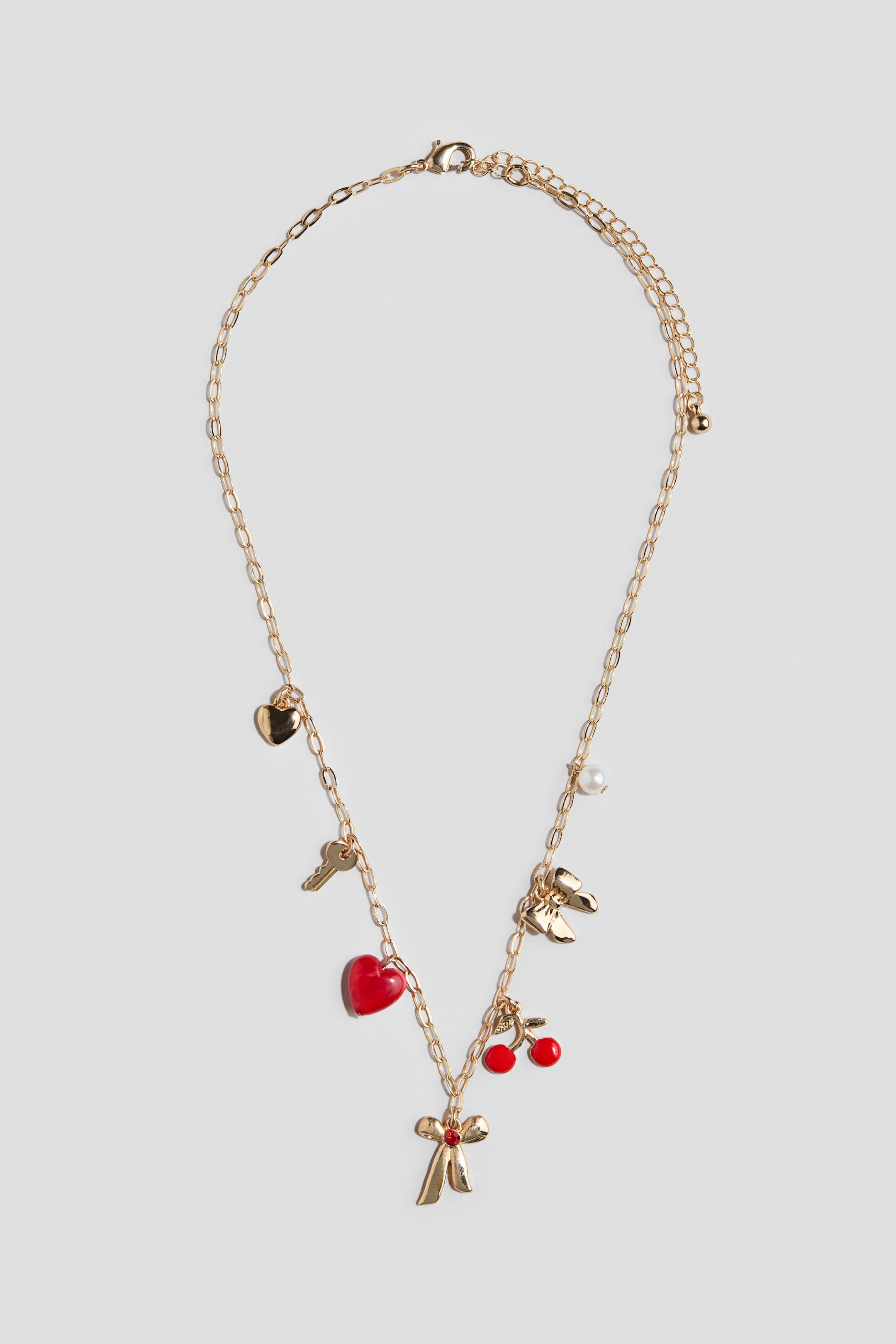 View larger image: A gold-toned chain necklace is displaying various charms along its length, including a small gold heart, a gold key, a red heart, a large gold bow with a red gem, red cherries, a pair of gold wings, and a white pearl. The necklace features a lobster clasp and an extender chain at the back.