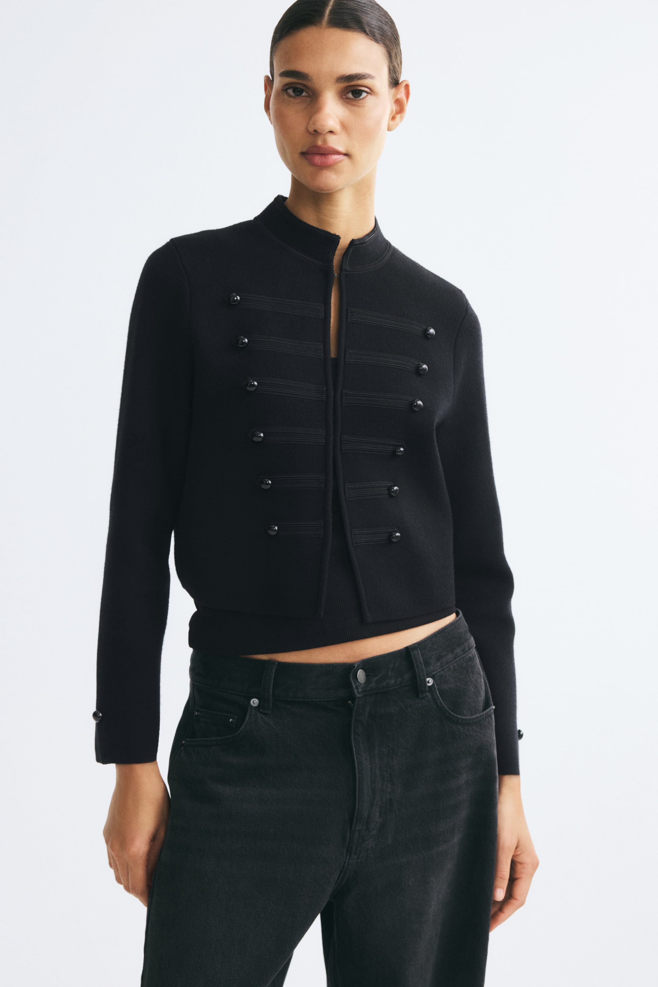 View larger image: A black, short knit cardigan featuring a stand collar, decorative front tabs with buttons, and long sleeves with buttoned cuffs is worn by the model. The open-front cardigan has a straight-cut hem and is styled with black jeans.