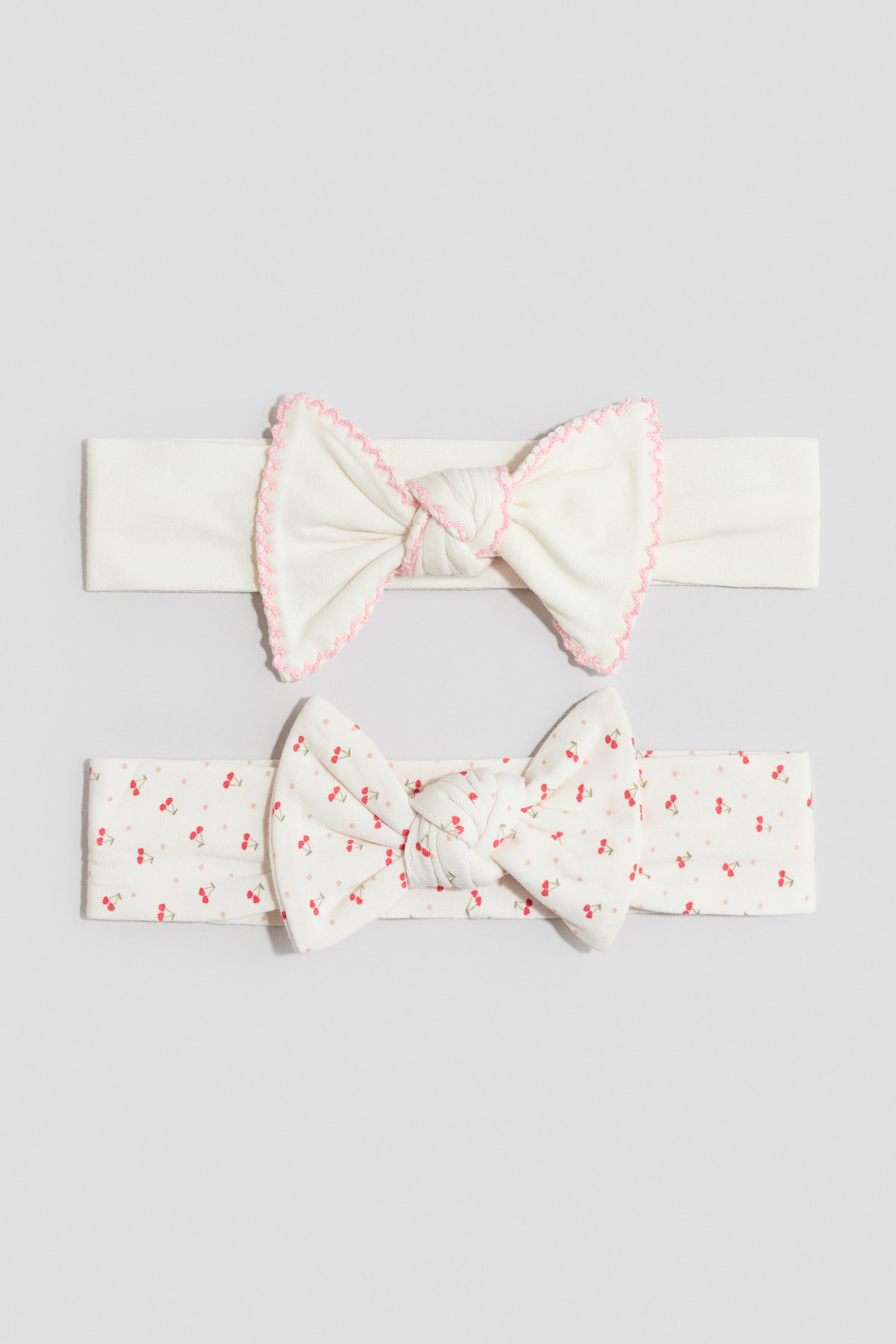 Größeres Bild ansehen: Zwei Jersey-Haarbänder präsentieren ihre Vorderseite. Das obere Haarband ist cremefarben und hat vorne eine Schleife mit rosa gewelltem Besatz entlang der Kanten. Das untere Haarband ist cremefarben mit einem all-over Kirschdruck und einer Schleife vorne.