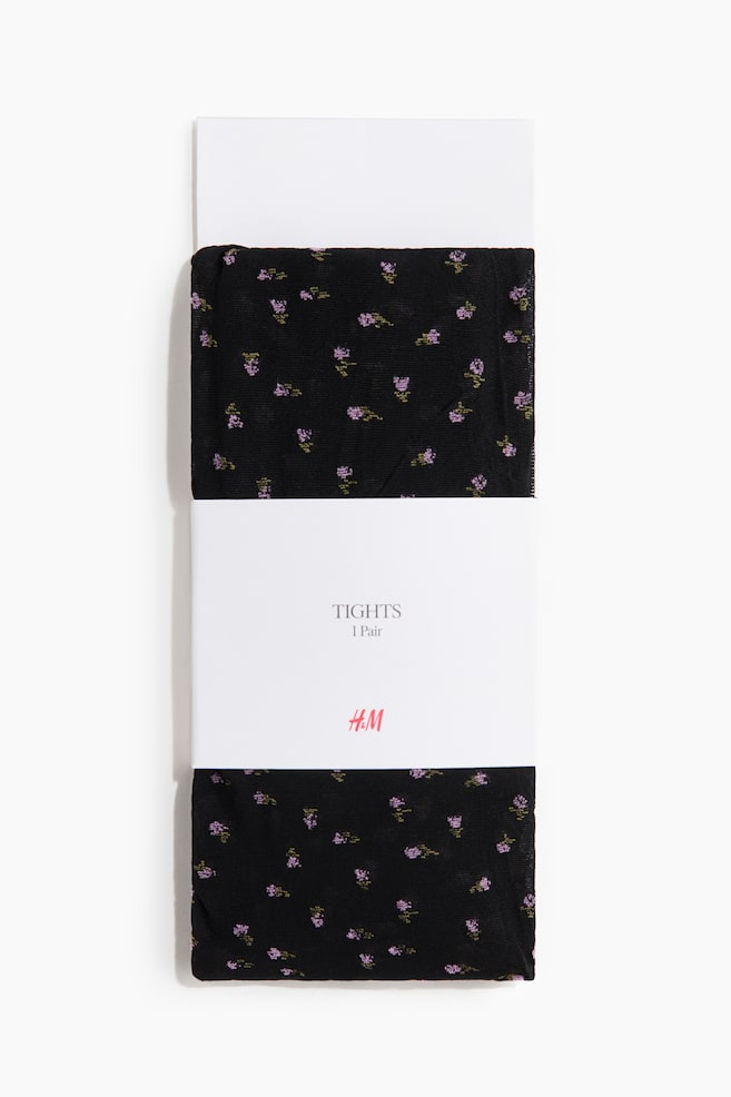 A pair of black tights featuring a small, scattered purple and green floral pattern is lying folded flat. A white paper band is wrapped around the centre of the tights, displaying the text "TIGHTS 1 Pair" and a pink logo. - 1