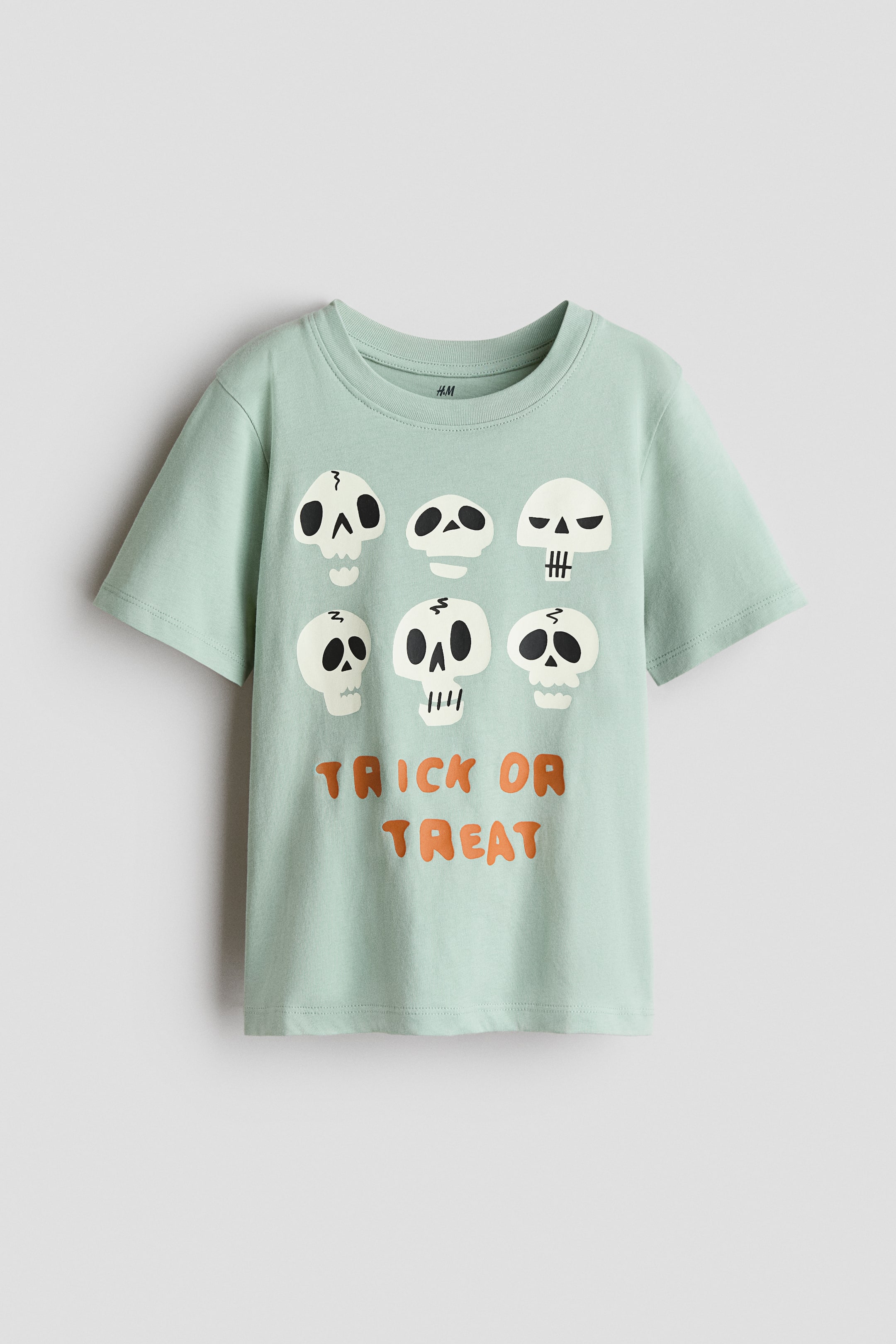 View larger image: A pale green short-sleeved t-shirt is hanging flat, facing forward. It features a ribbed crew neck and a graphic with six white cartoon skulls with black details across the chest, with the words 'TRICK OR TREAT' in orange bubble letters printed below the skulls.