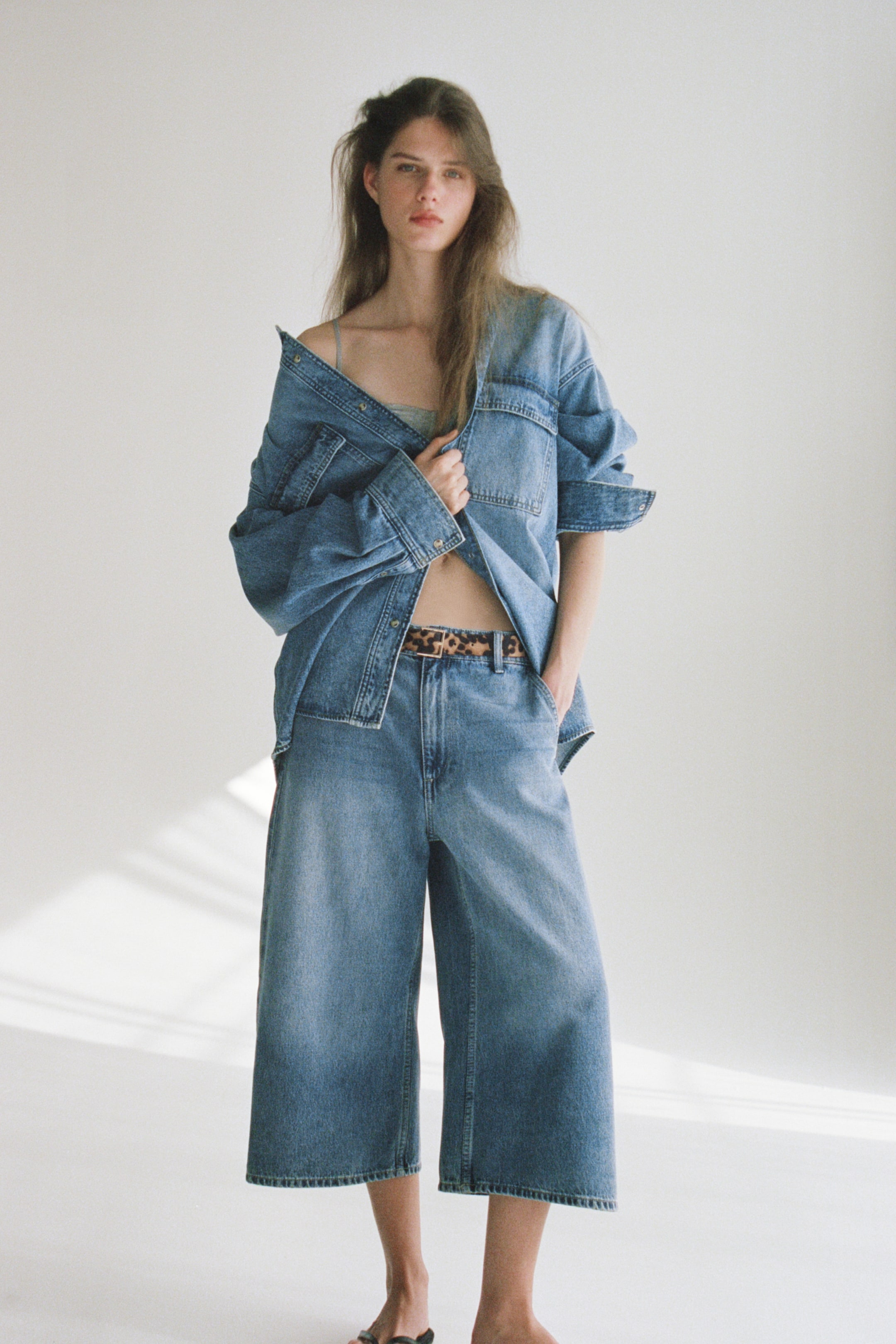 View larger image: Presenting a medium-wash denim set, showcasing an oversized shirt worn open with rolled-up sleeves and two chest pockets. Pair it with high-waisted, wide-leg culottes, cinched with a leopard print belt, and a light green bralette peeking beneath the open shirt.