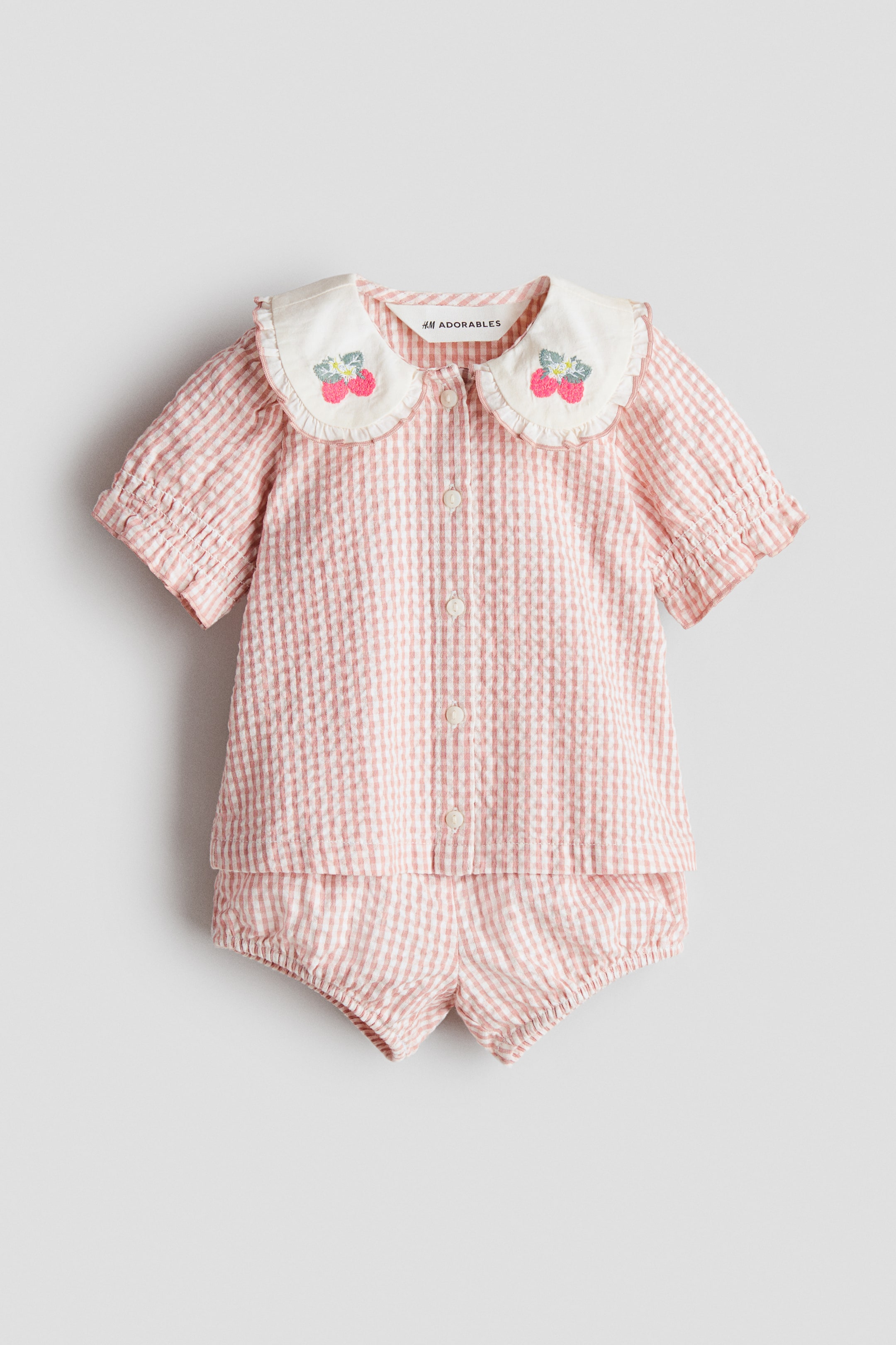 Größeres Bild ansehen: Ein zweiteiliges Baby-Set präsentiert in Vorderansicht ein rosa-weiß gestreiftes Seersucker-Shirt und passende Bloomer-Shorts. Das Shirt verfügt über eine durchgehende Knopfleiste, kurze Puffärmel mit elastischen Bündchen und einen weißen, gerüschten Peter-Pan-Kragen mit eingestickten roten Erdbeeren. Die Bloomer-Shorts vervollständigen das Set mit elastischen Beinabschlüssen.