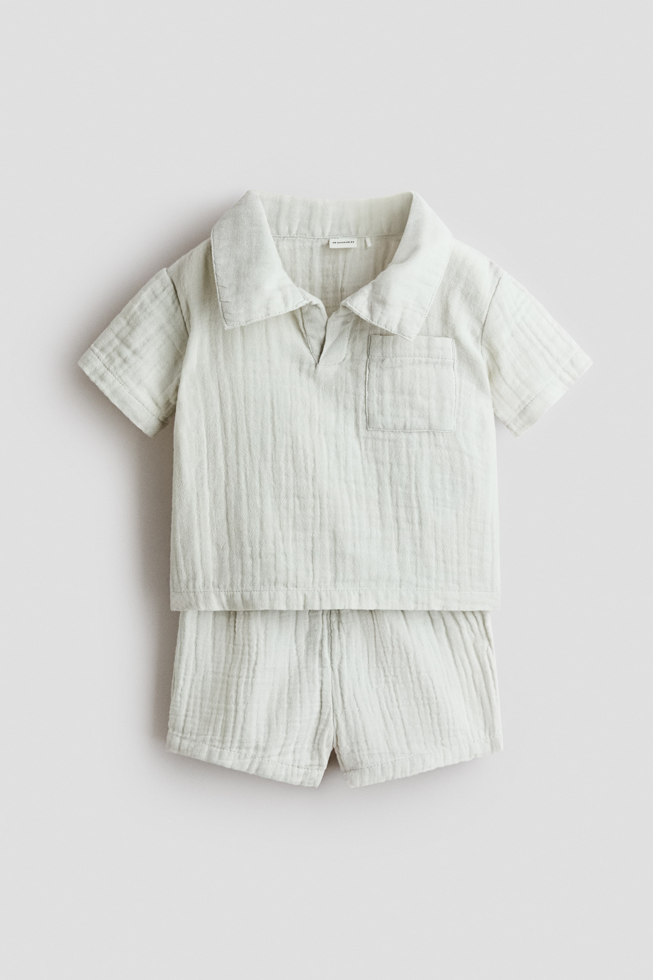 查看大圖: 兩件式嬰兒服裝套裝，正面展示，包含短袖上衣和配套短褲，均由皺褶的淺色面料製成。上衣設計有展開式領口、V字領開口，以及左胸的貼袋，而短褲則呈現寬鬆版型。