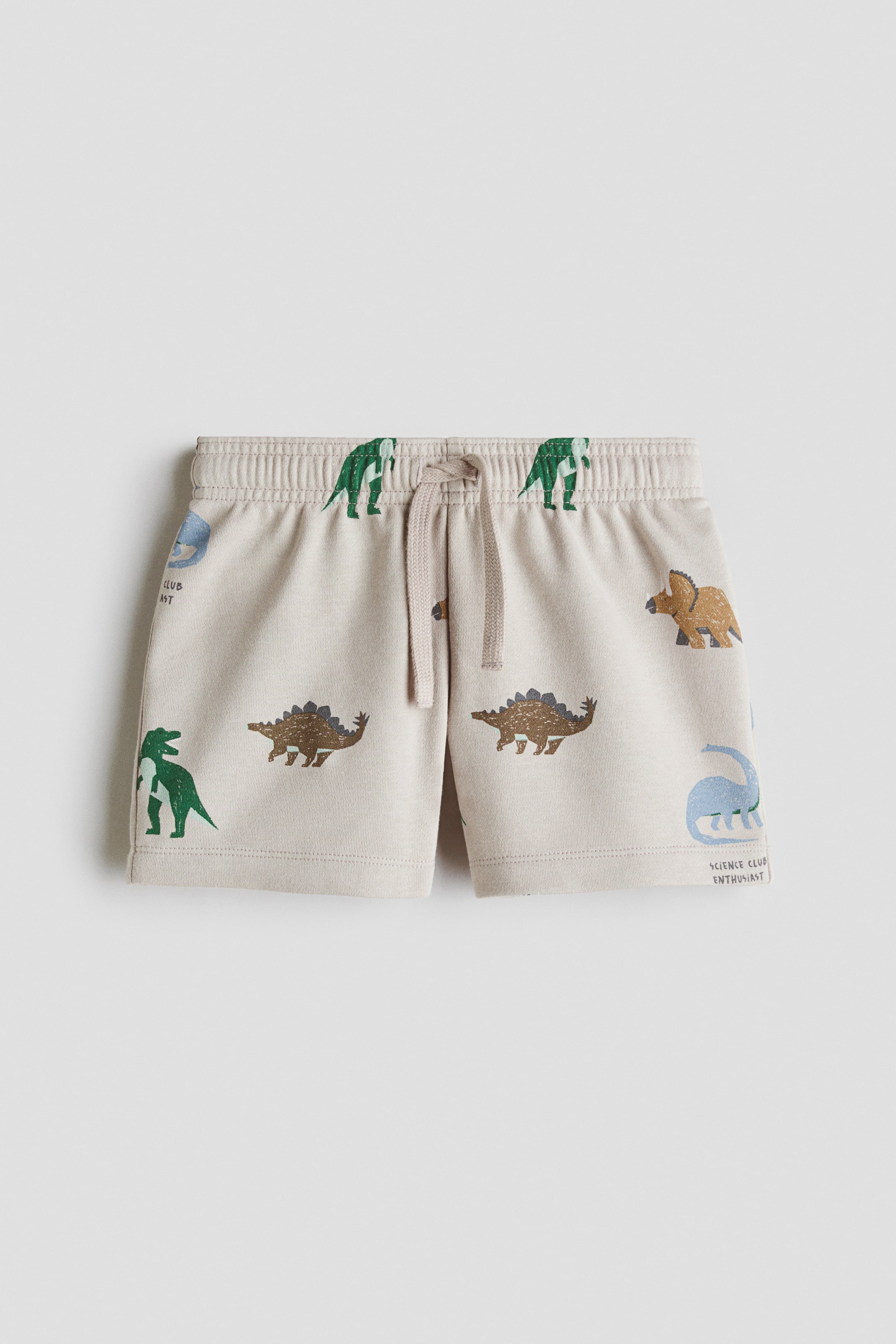 View larger image: The light beige shorts are shown in a front view, highlighting an elasticated ribbed waistband with a beige drawstring. The fabric is adorned with illustrations of dinosaurs in shades of green, brown, and blue, accompanied by the text 'science club enthusiast.'