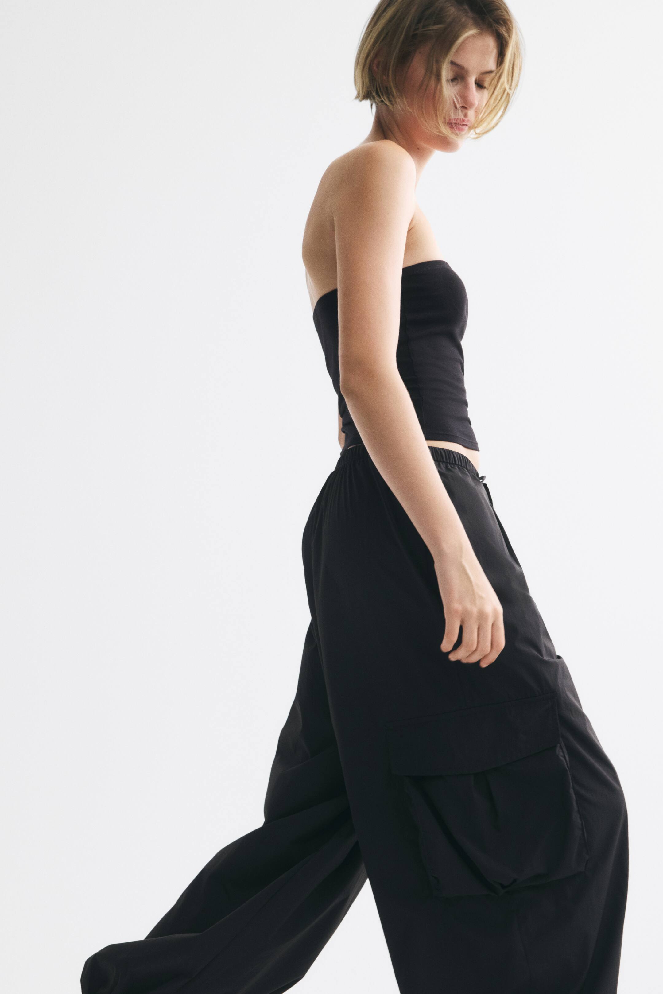 View larger image: The model is wearing oversized black nylon trousers with a strapless black top. The trousers feature an elasticated drawstring waist, wide balloon legs, bellows pockets with flaps, and elasticated hems.