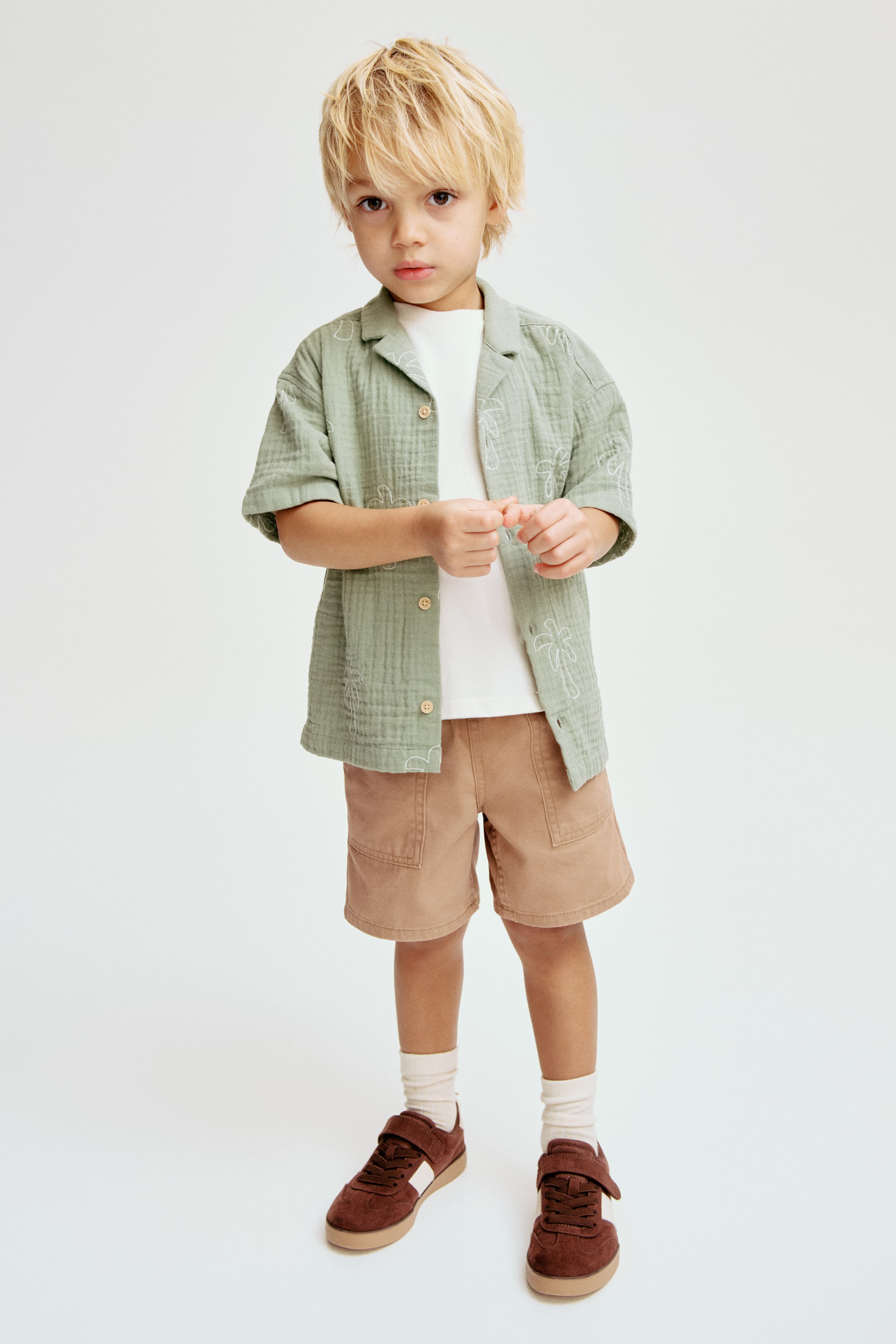 הצגת תמונה גדולה יותר: חולצה ירוקה בהירה עם שרוולים קצרים ורקמת עצי דקל לבנים נלבשת פתוחה מעל חולצת טי לבנה, בשילוב עם מכנסיים קצרים חומים. גרבי קרסול לבנים עם צלעות ונעלי ספורט חומות כהות עם פסים לבנים בצדדים וסגירות ולקרו משלימים את המראה.