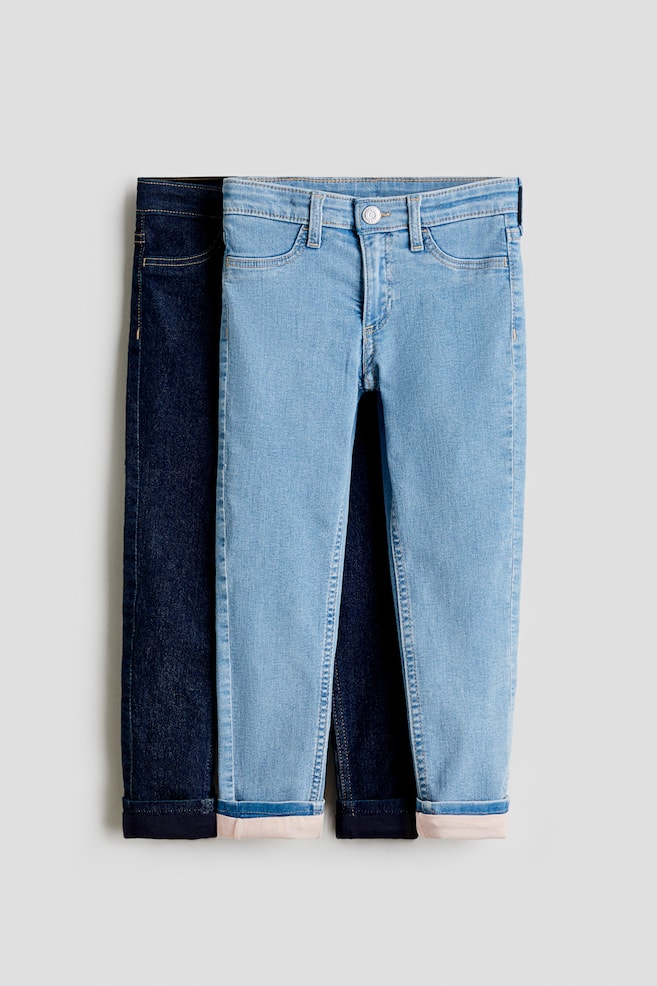 Zwei Paar eng anliegende, mit Jersey gefütterte Jeans aus dehnbarem Denim liegen flach, wobei das hellblaue Paar vorne positioniert ist. Das hellblaue Paar, das nach VORNE zeigt, verfügt über einen Reißverschluss mit Druckknopf, Gürtelschlaufen und falsche Vordertaschen, und das umgeschlagene Bein zeigt ein helles Jerseyfutter. - 1