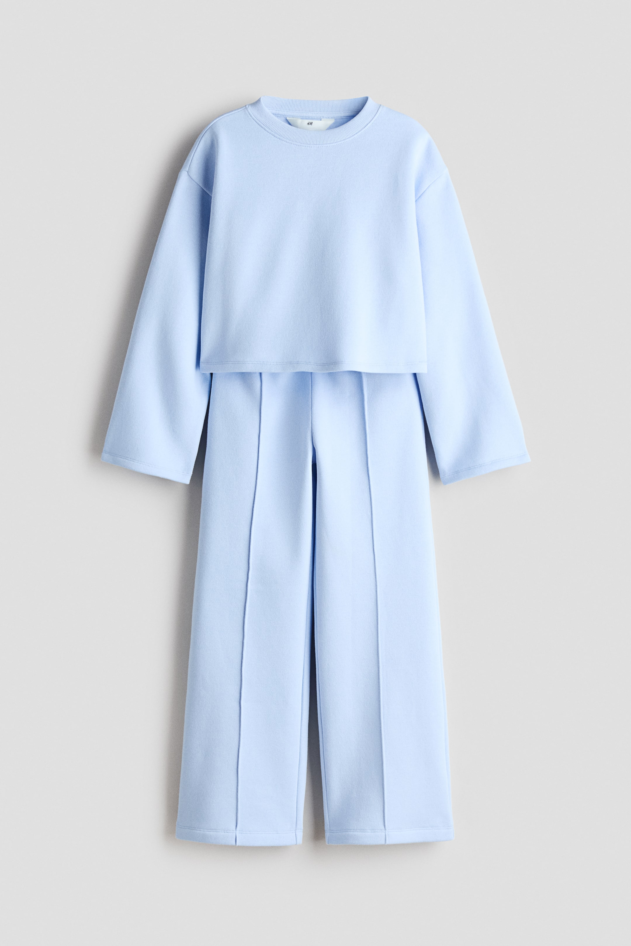 View larger image: A light blue sweatshirt set is showcased at the FRONT, featuring a long-sleeved top and matching joggers. The top boasts a round, ribbed neckline and dropped shoulders, while the joggers exhibit straight legs with sewn-in creases down the front.