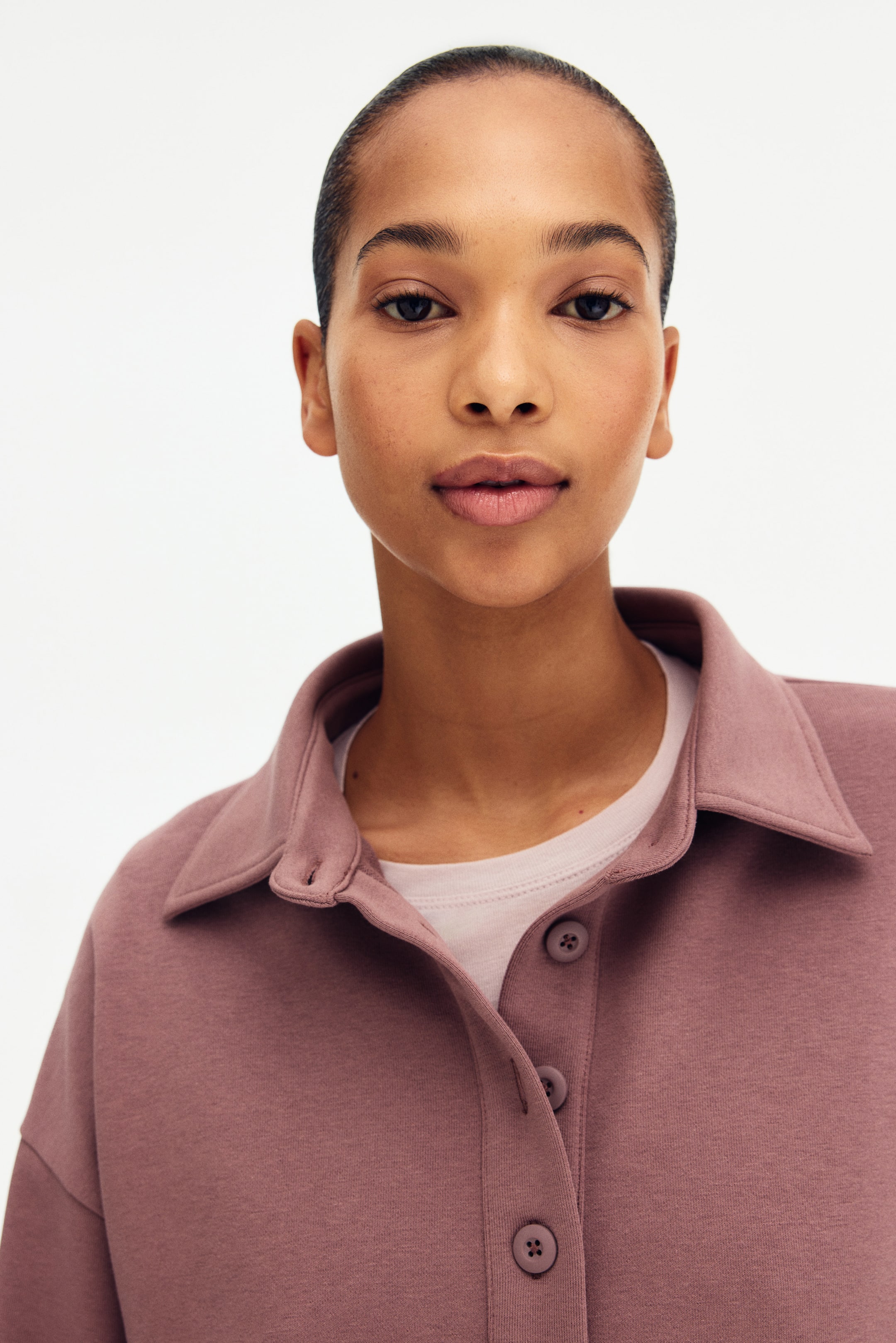 Grösseres Bild ansehen: Ein Sweatshirt in Altrosa mit klassischem Kragen und einer offenen Knopfleiste mit passenden runden Knöpfen, das über einem hellrosa T-Shirt getragen wird.