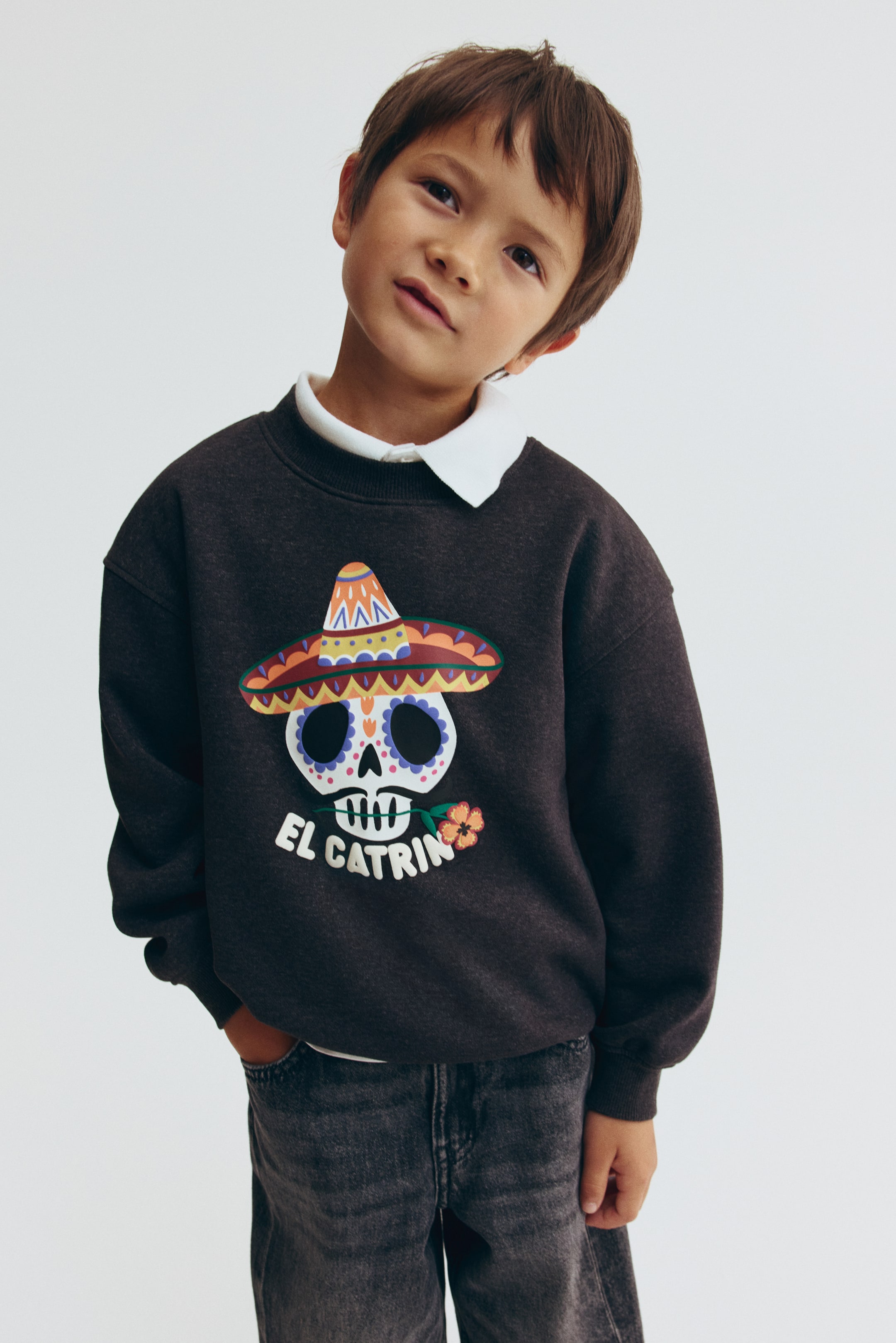 View larger image: A dark gray brushed fleece sweater is shown from the front, featuring a round rib-trimmed neckline, dropped shoulders, and ribbed cuffs and hem. The sweater features a colorful graphic print of a decorated skull wearing a sombrero with "EL CATR�N" text underneath, and is layered over a white collared shirt.