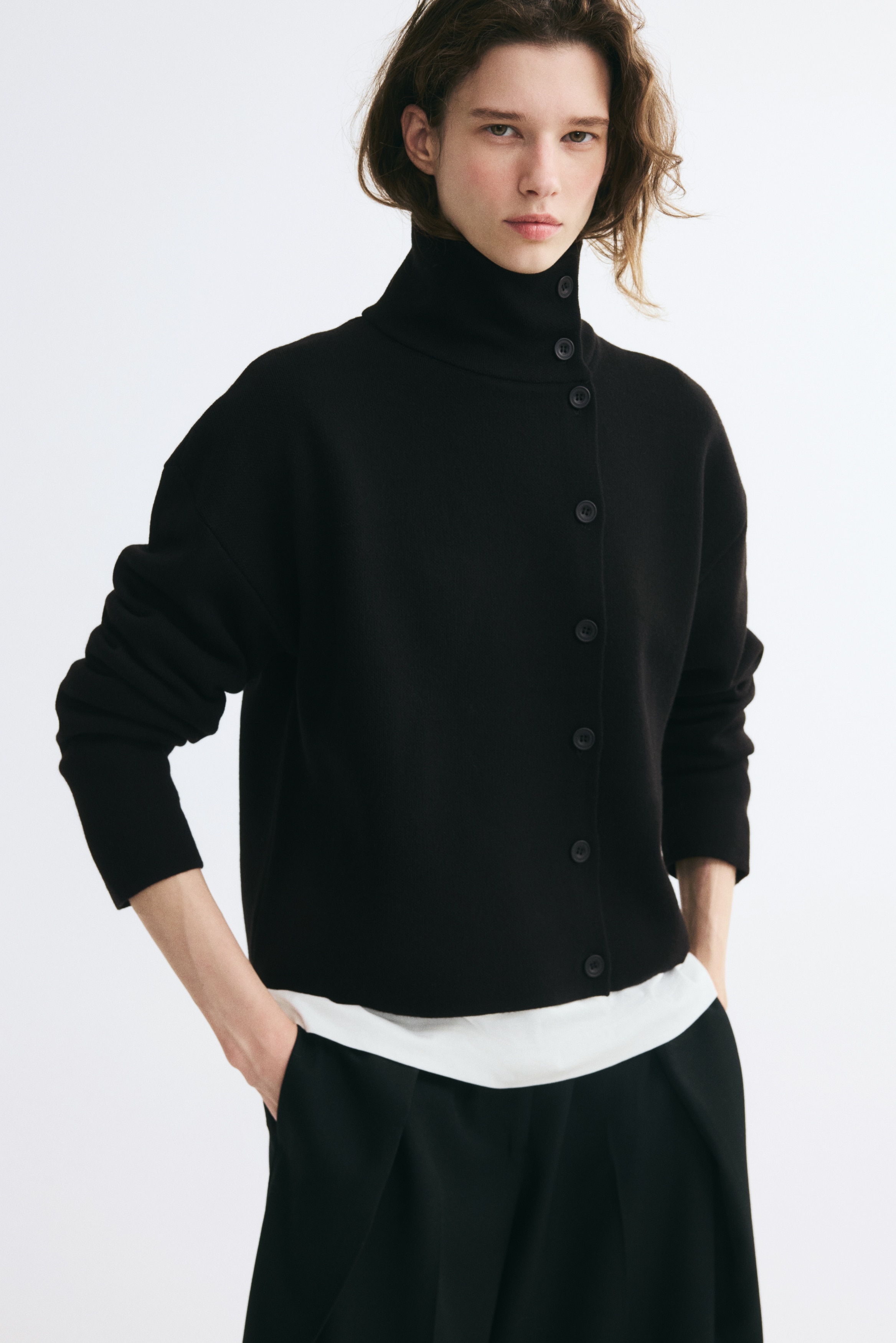 Un cardigan lavorato a maglia nero è mostrato frontalmente, con dolcevita alto e chiusura asimmetrica con bottoni sfalsata lungo il lato sinistro. Il capo è realizzato con spalle scese e maniche lunghe dritte in un morbido materiale a maglia fine.