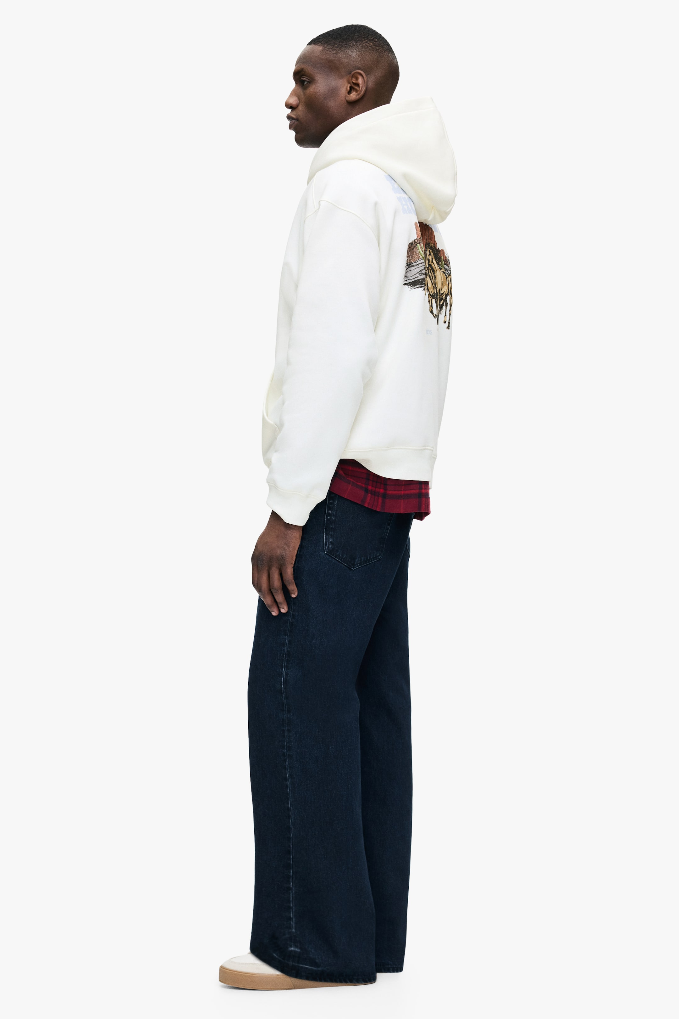 View larger image: A cream hoodie, featuring a colorful graphic print of a horse and landscape on the back and a front kangaroo pocket, is being worn by the model. A red plaid shirt is being layered underneath, completing the look with dark wash jeans and light-colored shoes.