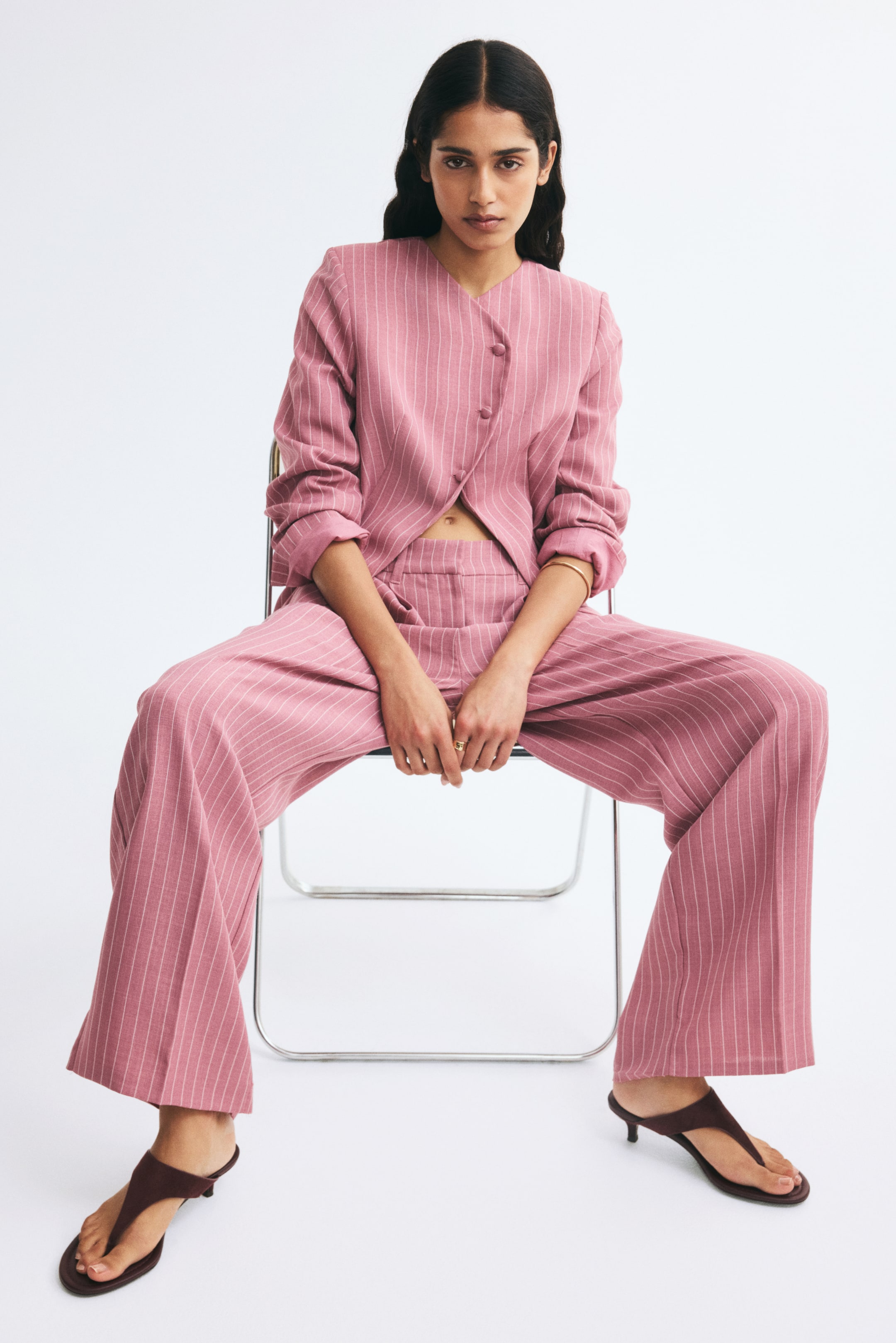 View larger image: These pink pinstriped woven trousers are shown from the front on a seated model, featuring a high-waisted fit with subtle front pleats and pressed creases running down the straight legs. The trousers are styled with a matching pink pinstriped long-sleeve top.