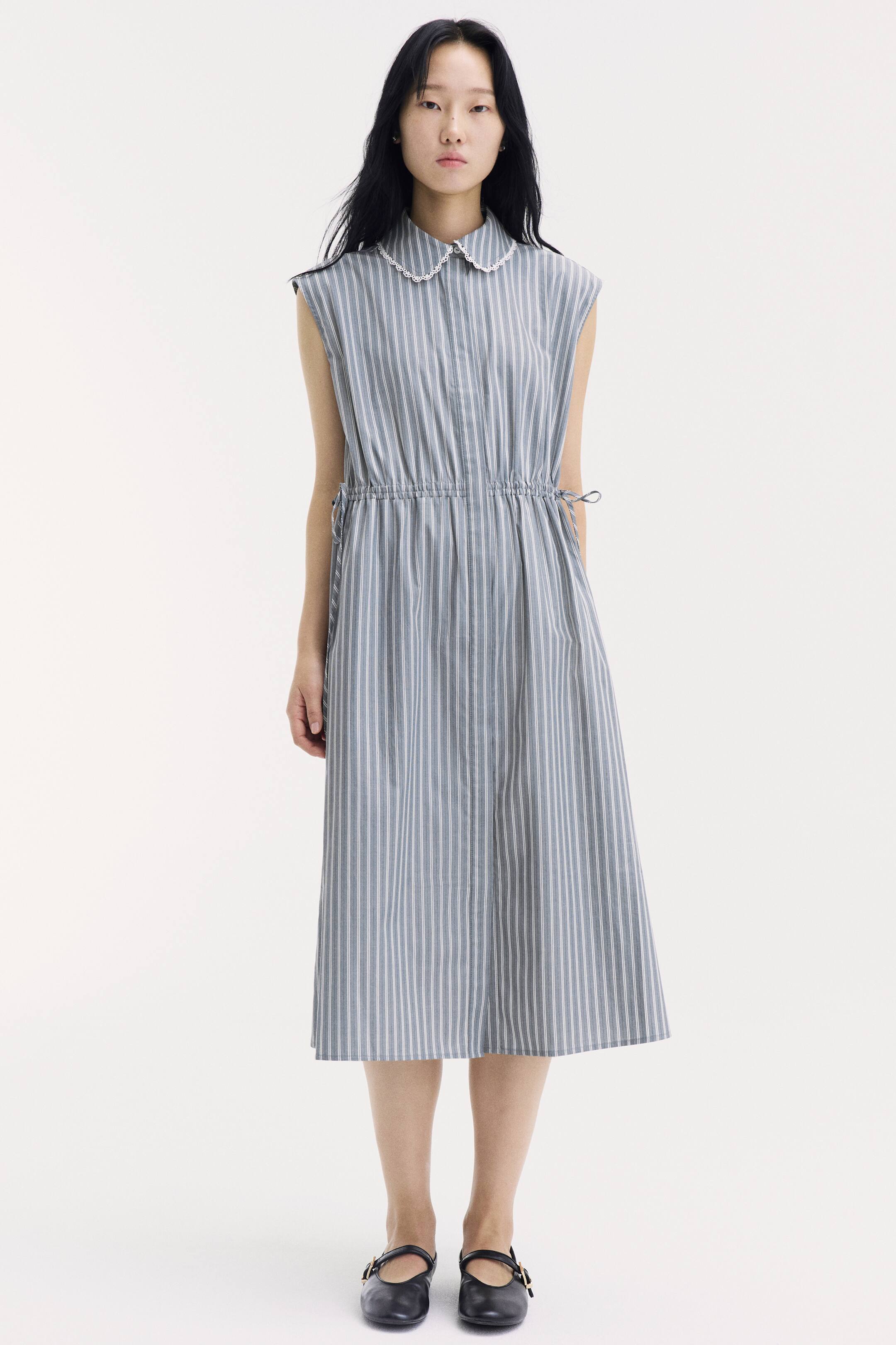 View larger image: A calf-length, sleeveless dress in a blue and white vertical stripe pattern is being worn by the model. The dress features a white lace-trimmed collar, a drawstring waist tied at the sides, and a gently flared skirt styled with black flat shoes.
