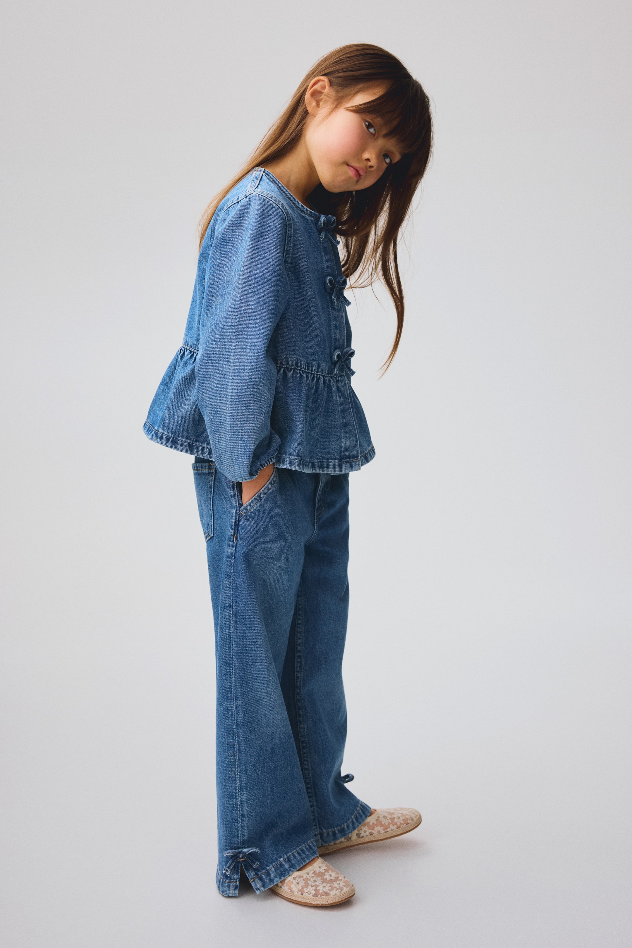 Visualizza immagine più grande: Un completo coordinato in denim per bambini è in mostra, composto da un top peplum con scollo rotondo e maniche lunghe, caratterizzato da diversi piccoli fiocchi in denim che scendono lungo la parte anteriore. I pantaloni in denim a gamba larga abbinati completano il set, includendo tasche laterali e orli con spacchi adornati con piccoli fiocchi in denim alle caviglie.