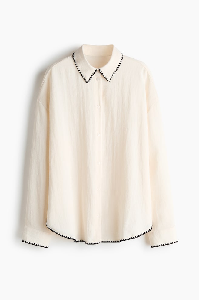 A cream-colored shirt is showcasing its front, featuring a pointed collar and a full button placket with light buttons. The collar, buttoned cuffs, and curved hem are all edged with a black scalloped trim. - 1