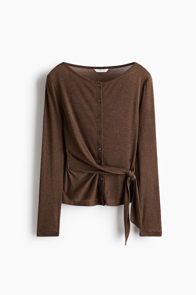 A brown knit top is shown front-facing, featuring a delicate boat neckline, long sleeves, and a button-down front. A triangular fabric panel wraps across the waist from one side and fastens with a self-tie on the opposite side. - 1