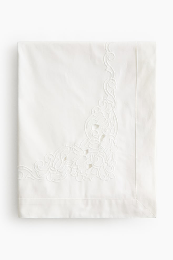 A white embroidered textile is lying flat, folded into a neat rectangle. Intricate white-on-white embroidery adorns one corner, showcasing swirling floral motifs and cutwork details. - 1