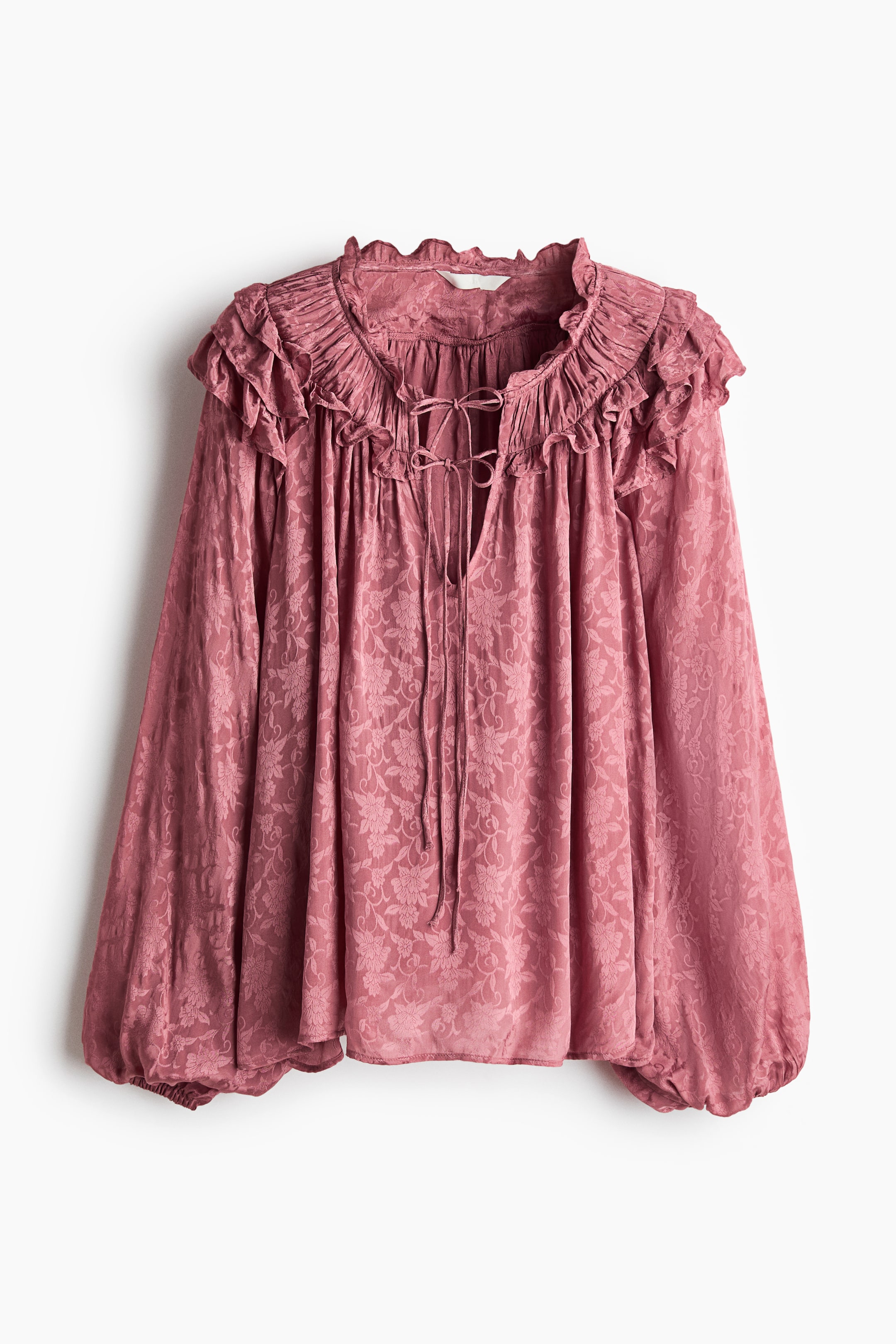 View larger image: A pink blouse features a subtle floral jacquard pattern throughout the fabric. It is designed with a ruffled high neck, a V-neck opening secured by matching ties, and voluminous long sleeves gathered into elasticated cuffs.