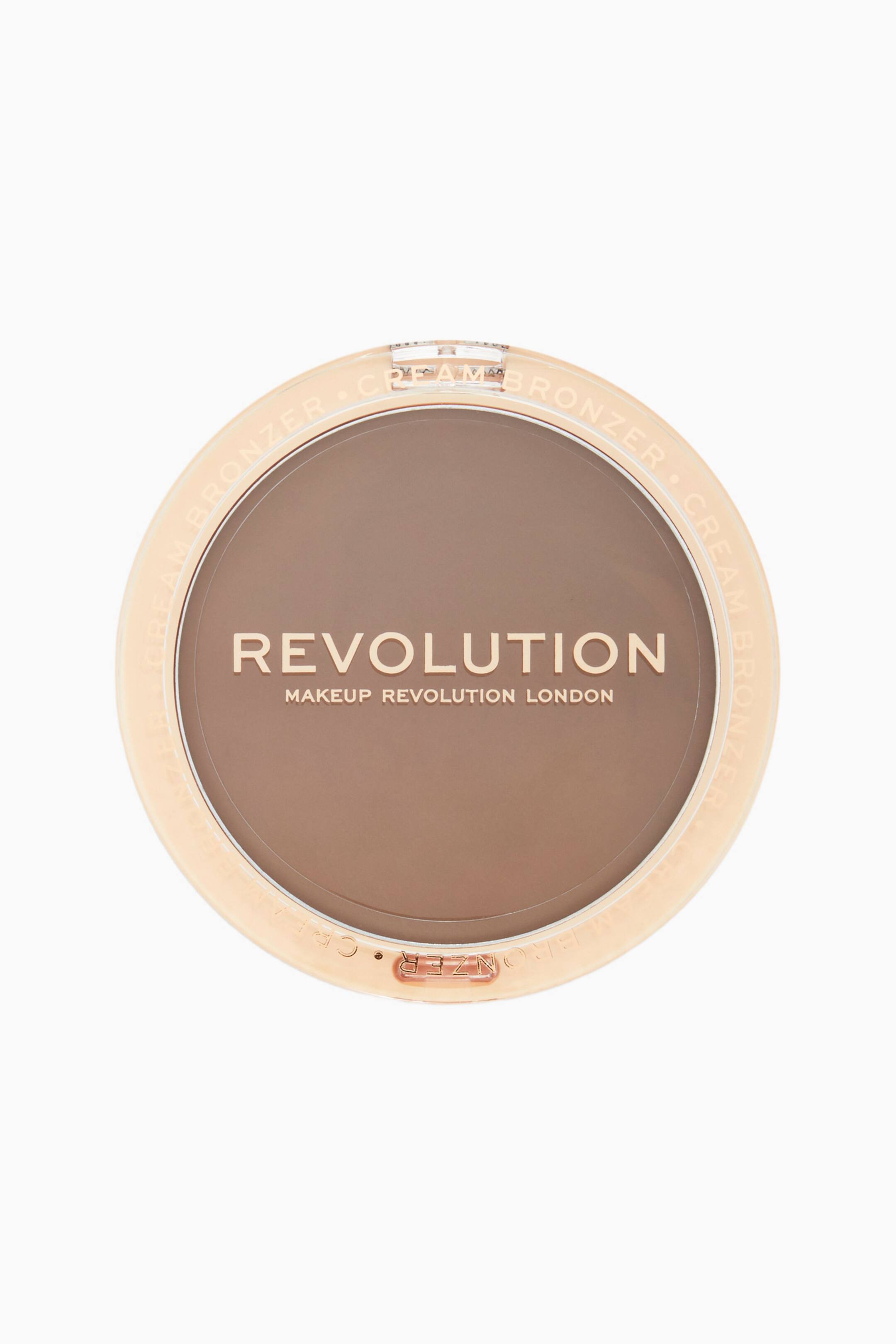 Se større billede: Et top-down billede viser Revolution Ultra Cream Bronzer i en rund, gennemsigtig plastikæske fyldt med en jævn, mediumbrun creme. Guldbogstaver viser brandnavnet "REVOLUTION" på tværs af midten, mens ordene "CREAM BRONZER" gentages langs lågets ydre, cirkulære kant.