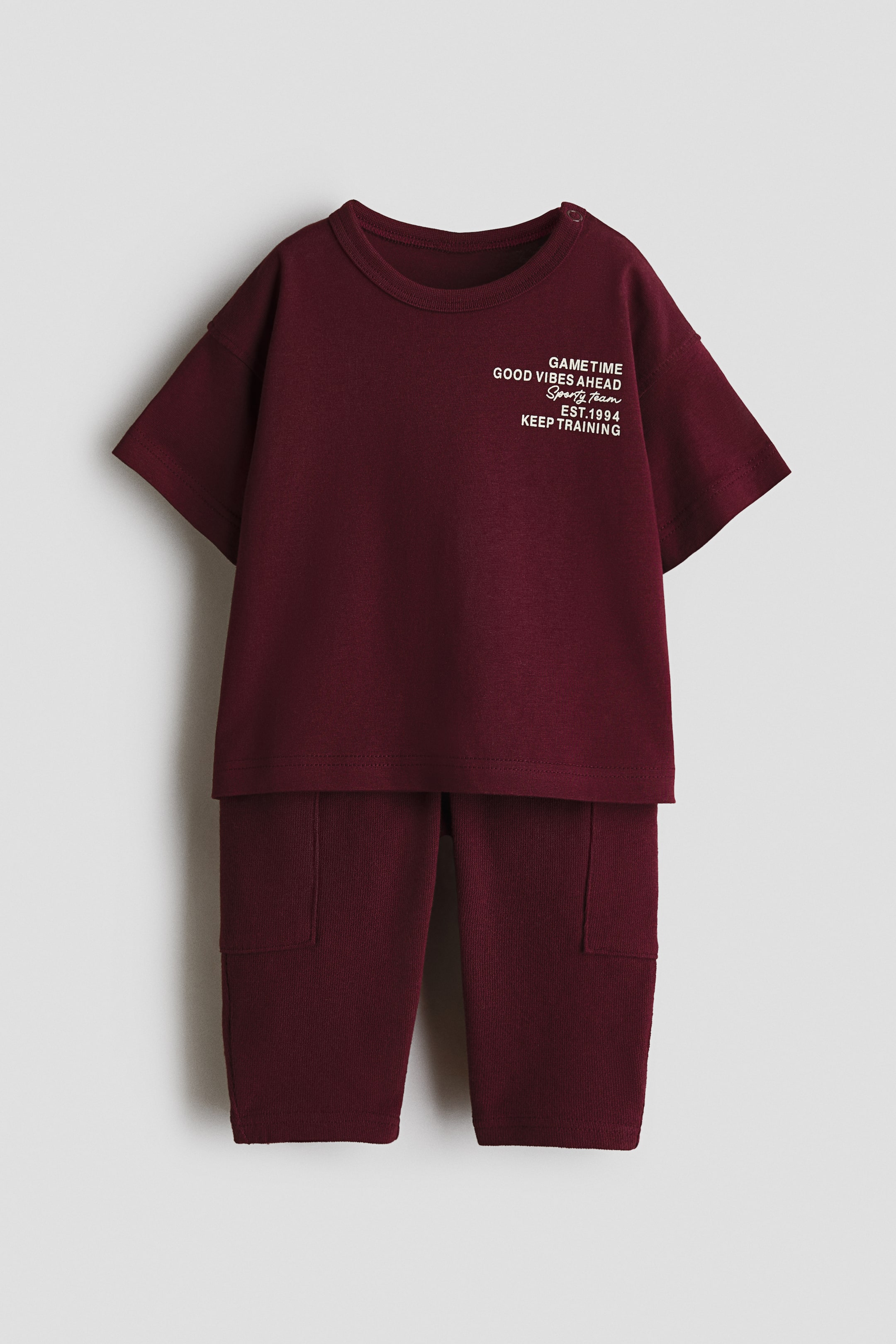View larger image: A front view of a maroon 2-piece cotton set features a short-sleeved T-shirt with a ribbed crew neck, white text graphic on the chest, and snap buttons on one shoulder, paired with long pants featuring two patch pockets on the thighs.