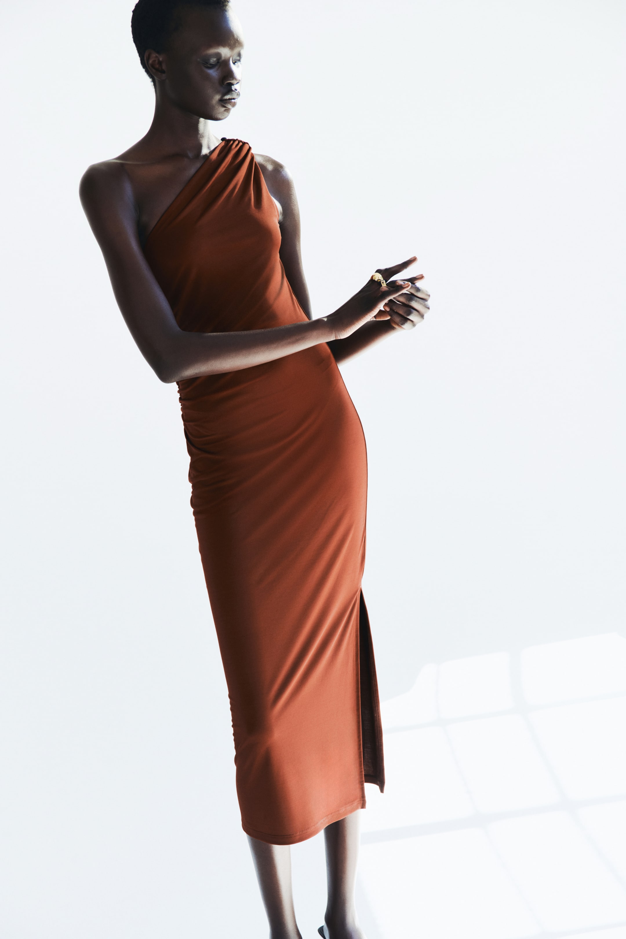 View larger image: A rust-colored one-shoulder midi dress is showcased from a slight side angle, highlighting its draped bodice and tailored silhouette. The dress is crafted from smooth fabric, with subtle ruching on the right side of the waist and a high slit on the right side of the skirt.
