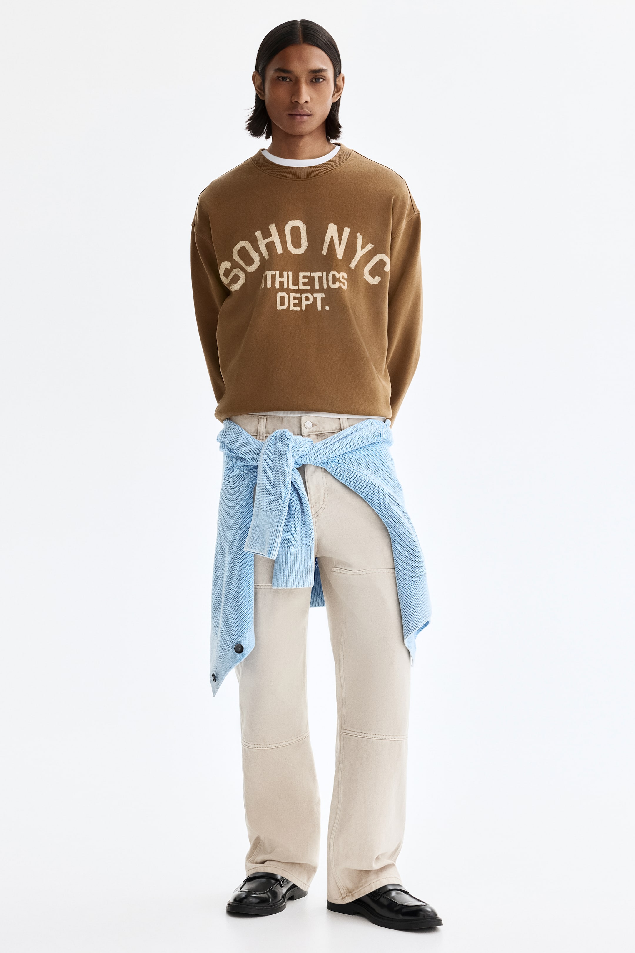 View larger image: A brown crewneck sweatshirt is shown front-facing, featuring "SOHO NYC ATHLETICS DEPT." in distressed, light-colored block letters across the front. It has long sleeves, ribbed cuffs, and a ribbed hem, with a white T-shirt layered under the crewneck.
