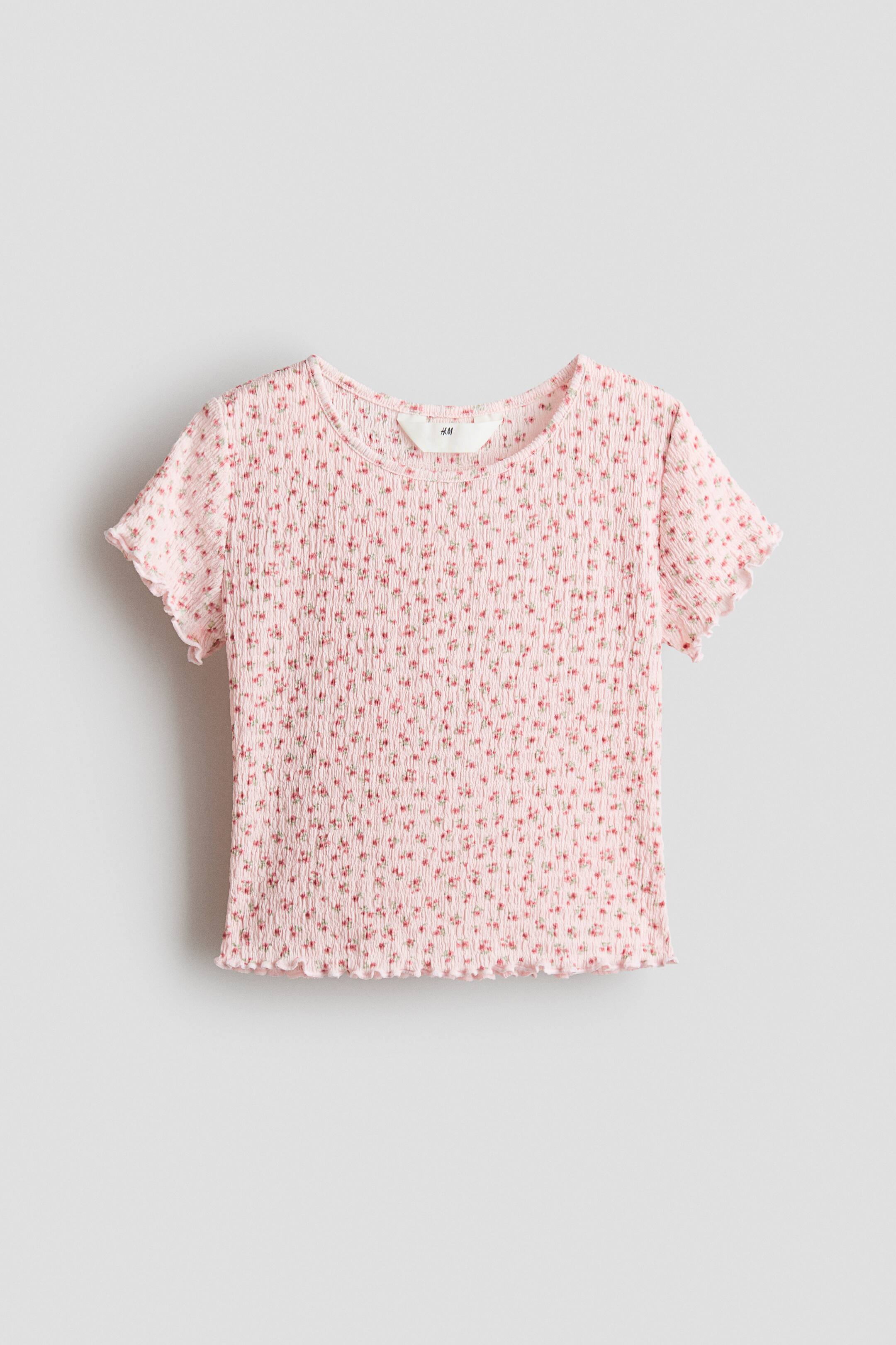 View larger image: A light-pink shirred short-sleeved top is shown from the front, featuring a small red-and-green floral pattern. It is designed with a round neckline and lettuce-edge hems on the sleeves and hem, with an H&M tag visible at the neckline.