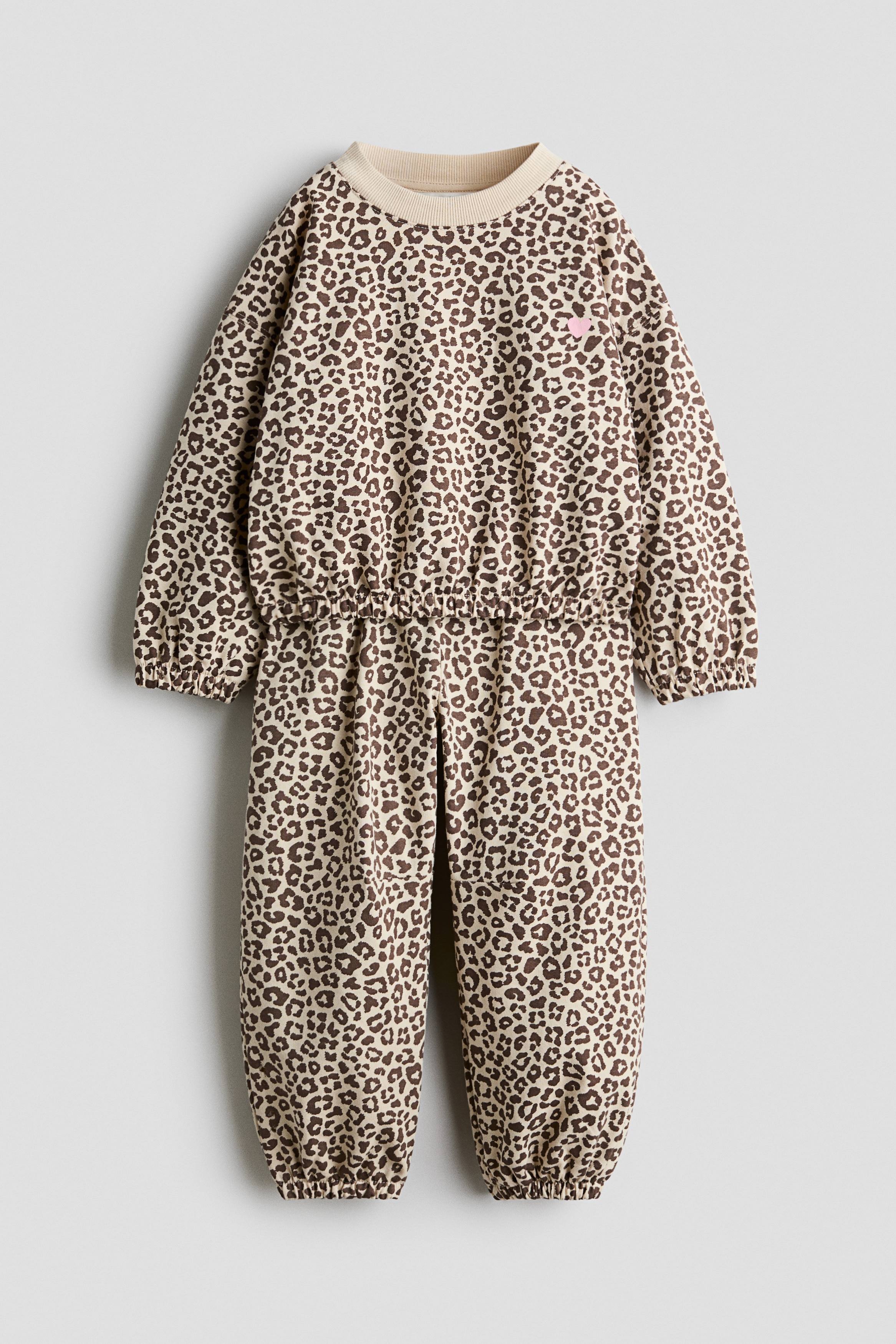 A front-facing matching leopard-print fleece set is shown, consisting of a long-sleeve sweater and relaxed track pants. The sweater features a cream ribbed crew neck, dropped shoulders, voluminous sleeves with burgundy ribbed cuffs, and a matching burgundy ribbed hem. The matching track pants drape in a relaxed fit with gathered elasticated hems.