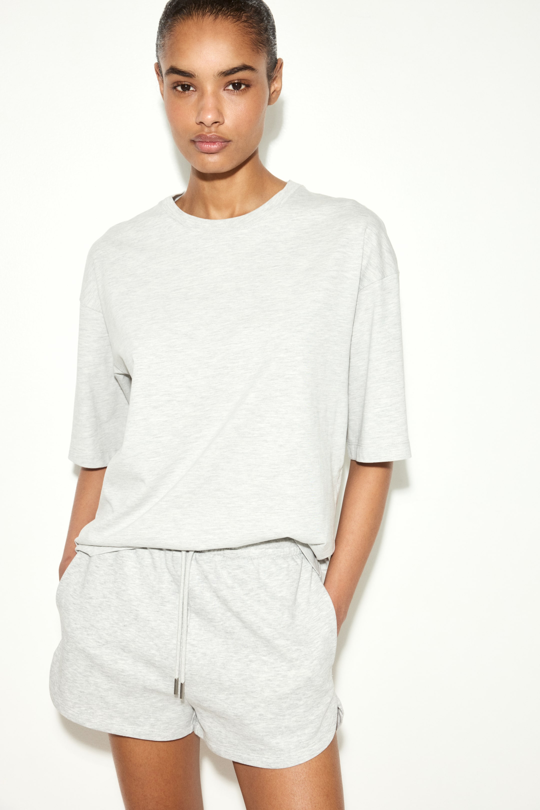 Ver imagen más grande: Se presenta una camiseta de cuello redondo en gris jaspeado con mangas cortas, combinada con unos pantalones cortos a juego en gris jaspeado que cuentan con una cintura ajustable con cordón y puntas metálicas, además de bolsillos laterales. Ambas prendas muestran un ajuste relajado.