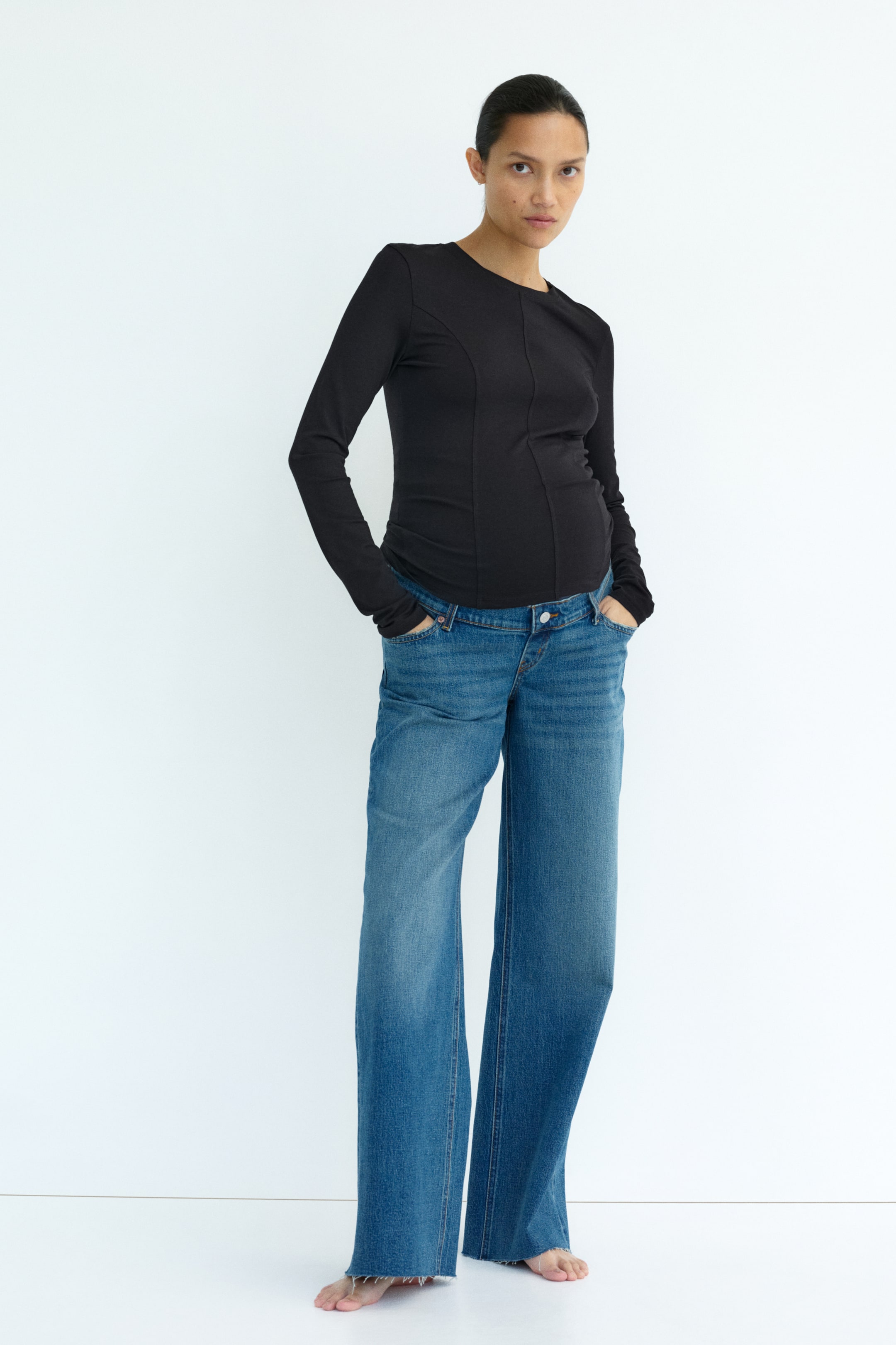 View larger image: The model is wearing five-pocket jeans in blue denim, with her hands placed in the front pockets. The jeans boast a relaxed fit, complete with a button and fly detail, and wide, straight legs. A wide jersey panel extends from the waist over the model's midsection.