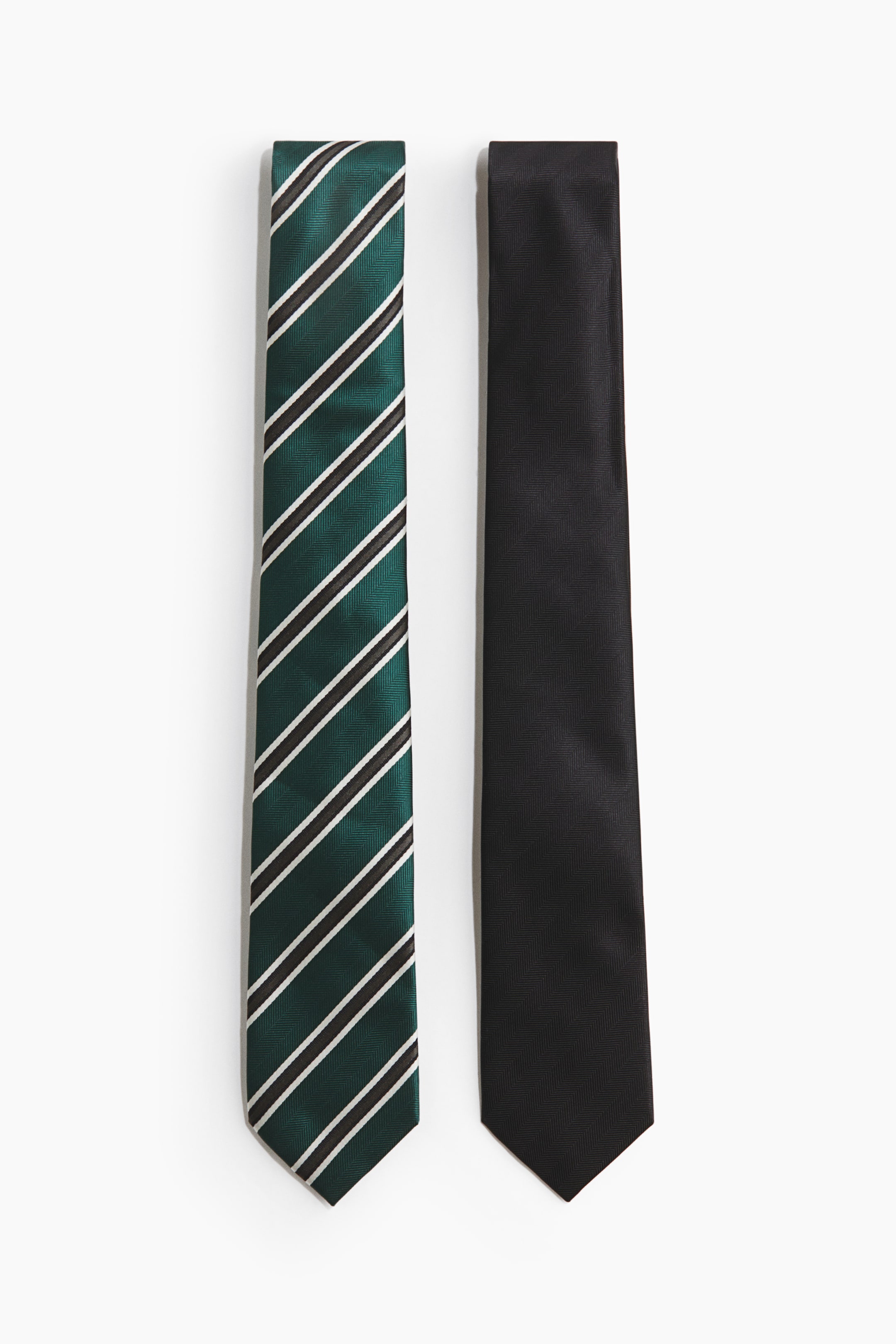 Ampliar la imagen: Dos corbatas están colocadas en posición horizontal. La corbata de la izquierda presenta un color verde oscuro con franjas diagonales negras y blancas y un sutil tejido espiga. La corbata de la derecha muestra un color negro sólido y un sutil tejido espiga.